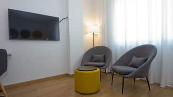 Una sala de estar moderna con un televisor de pantalla plana, dos sillones grises, un reposapiés amarillo y una lámpara de pie.
