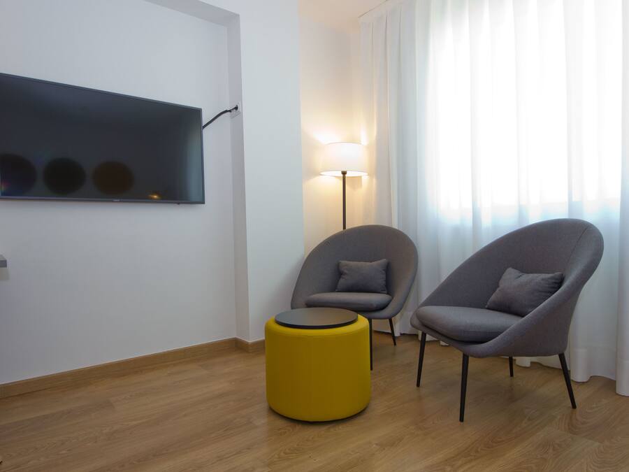 Una sala de estar moderna con un televisor de pantalla plana, dos sillones grises, un reposapiés amarillo y una lámpara de pie.