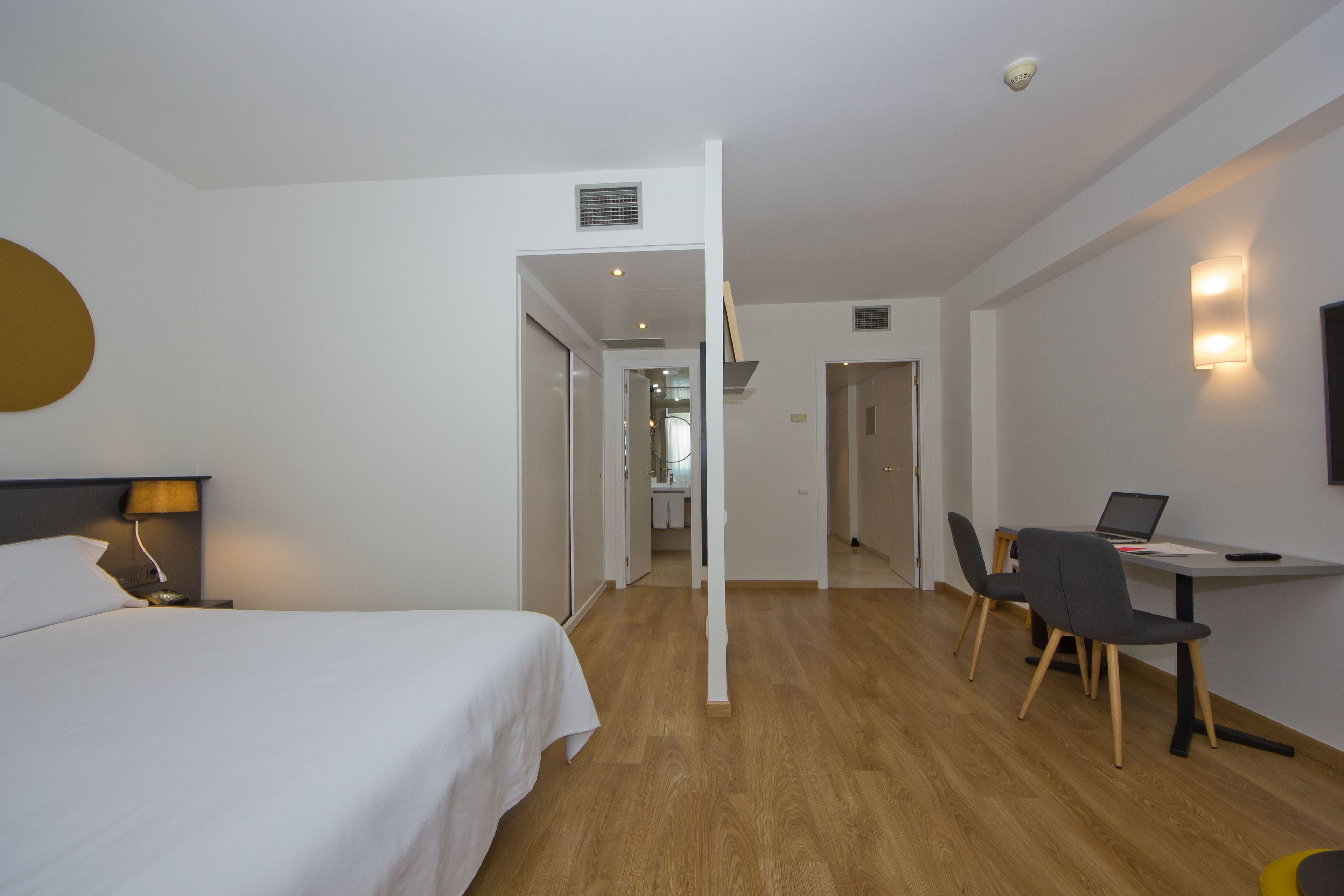 A hotel room with a bed, desk, and chair, a bathroom in the background, and a hallway leading to another room.