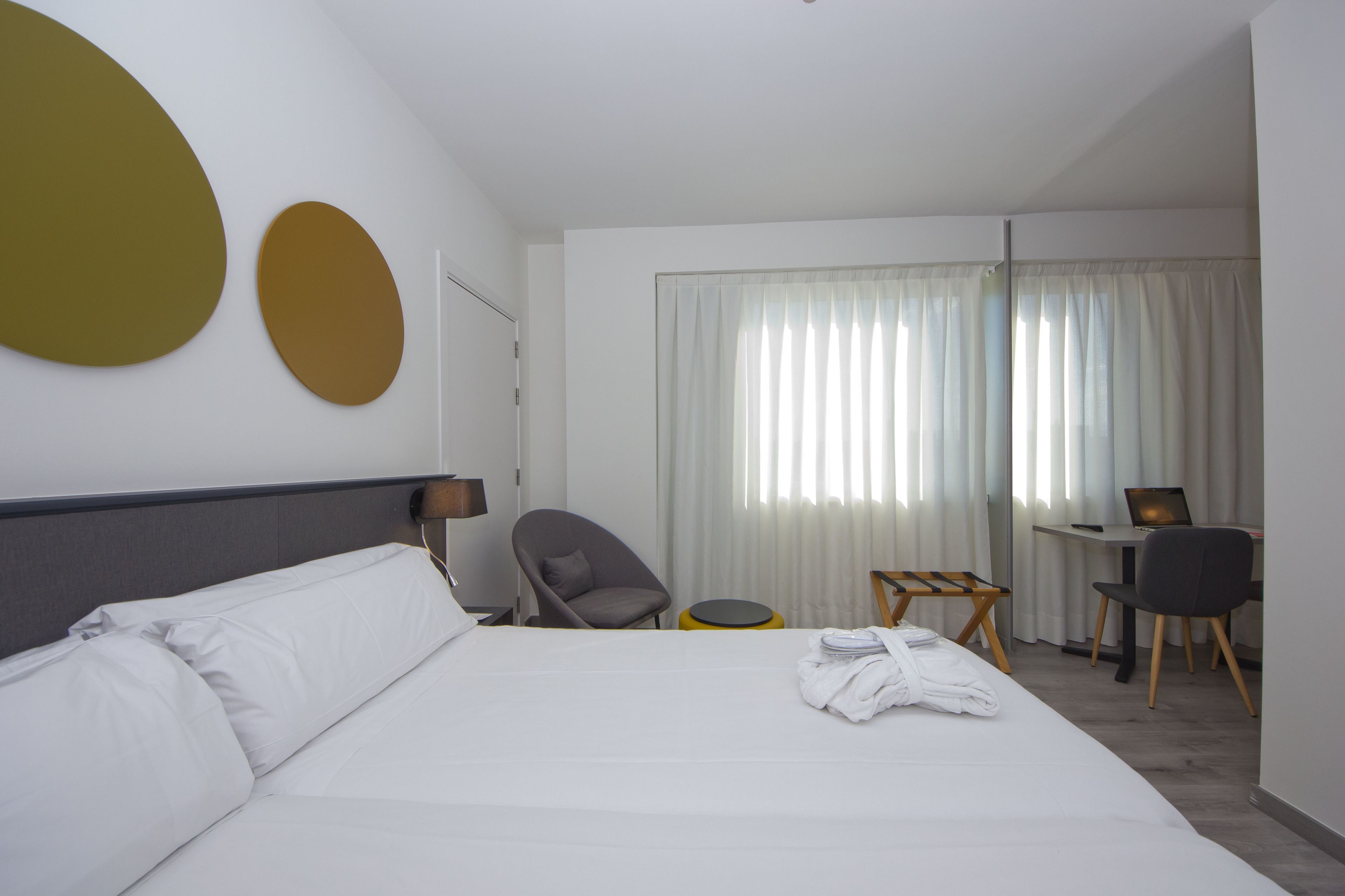 Une chambre d’hôtel avec un lit, un bureau, deux chaises, une petite table ronde et une fenêtre avec des rideaux.