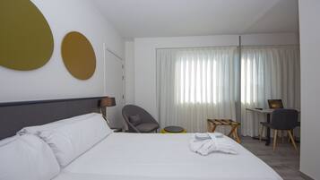 Une chambre d’hôtel avec un lit, un bureau, deux chaises, une petite table ronde et une fenêtre avec des rideaux.