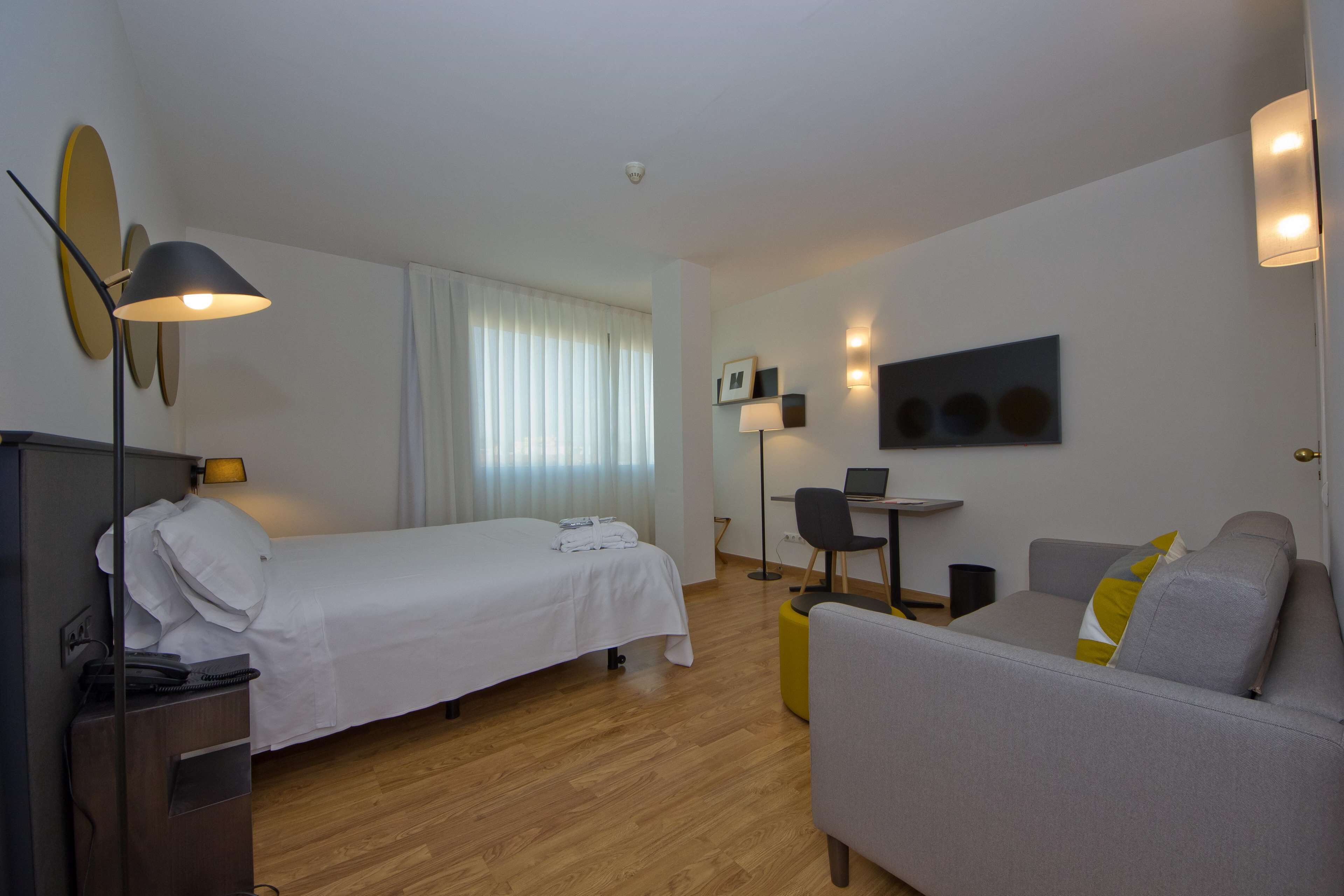 A hotel room with a bed, a desk with a chair, a sofa, and a lamp.