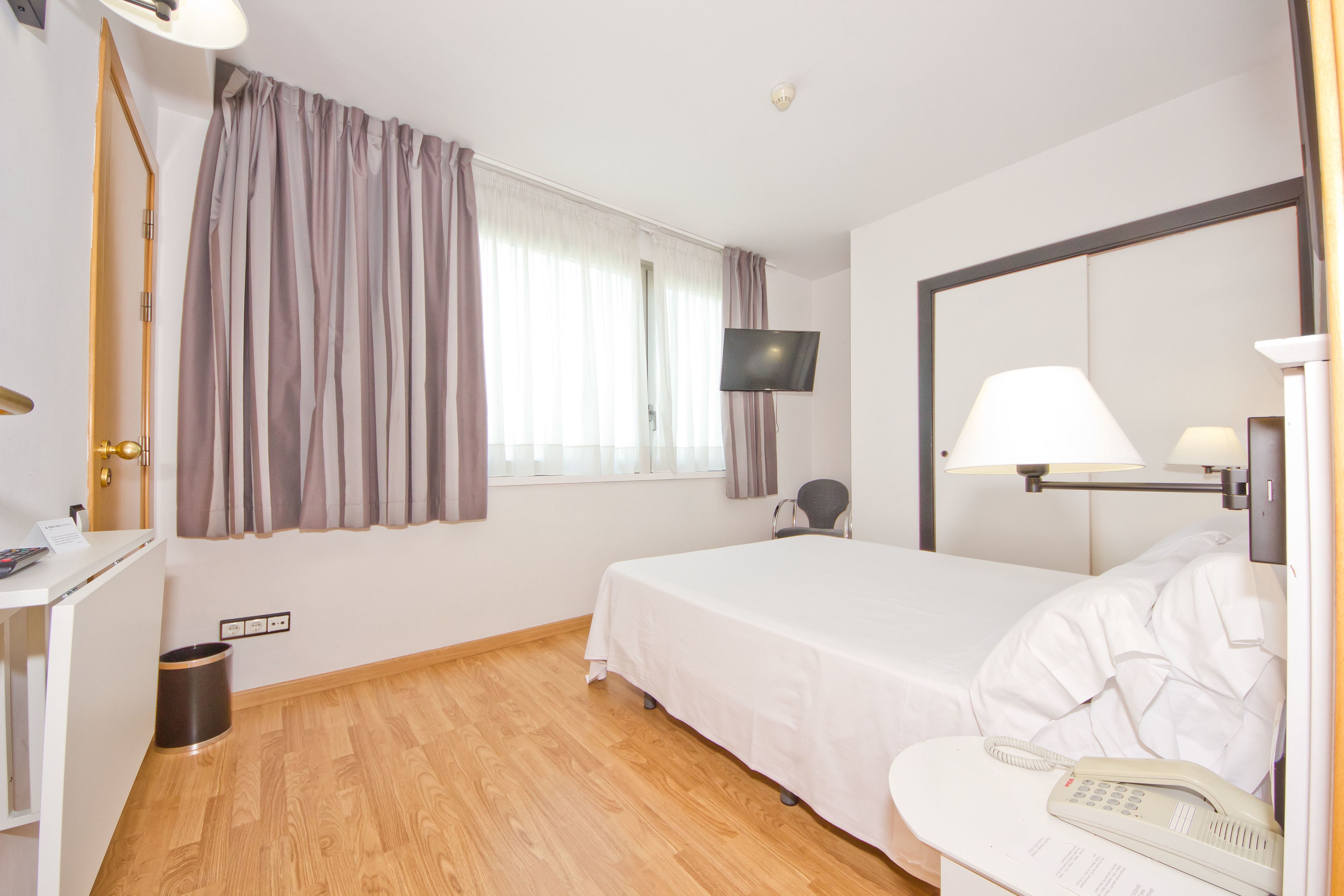 Une chambre d’hôtel avec un lit, un bureau avec une lampe, un téléphone, un téléviseur fixé au mur et une fenêtre avec des rideaux.