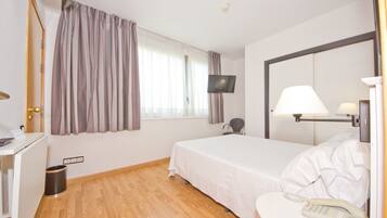 Habitación de hotel con cama, escritorio con lámpara, teléfono, televisor montado en la pared y ventana con cortinas.