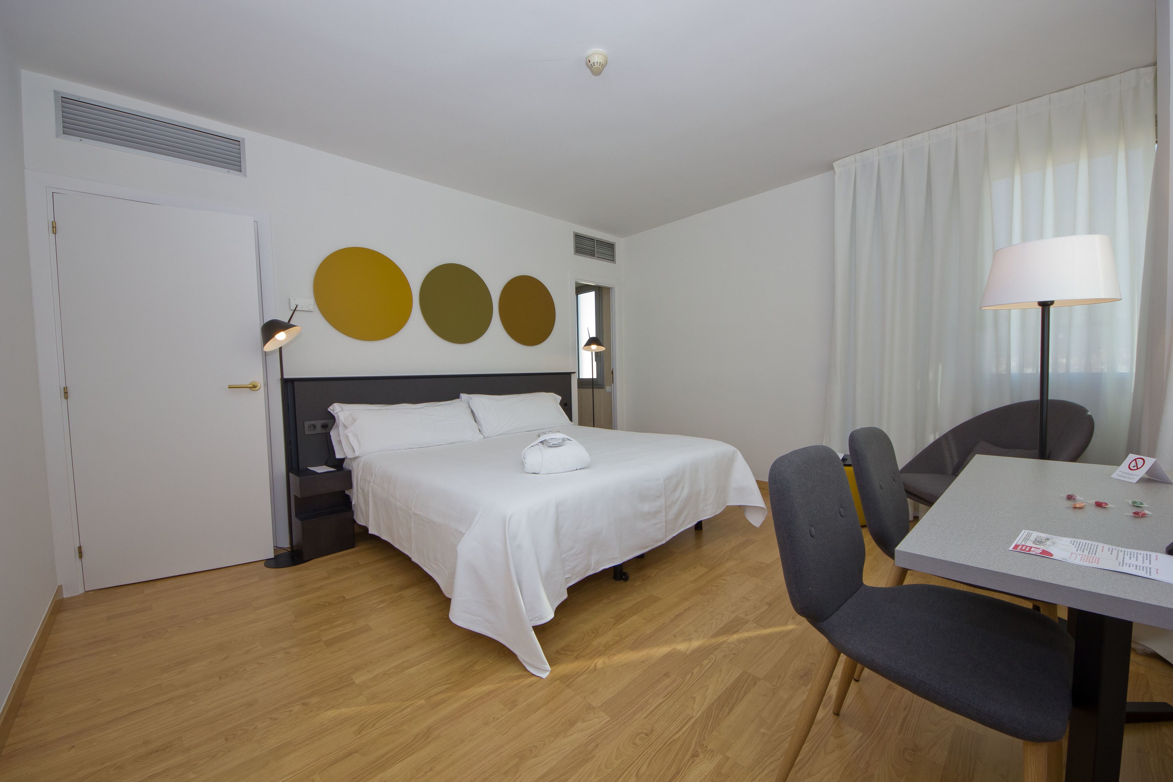 Een hotelkamer met een bed, nachtkastje, lamp en een eettafel met stoelen.