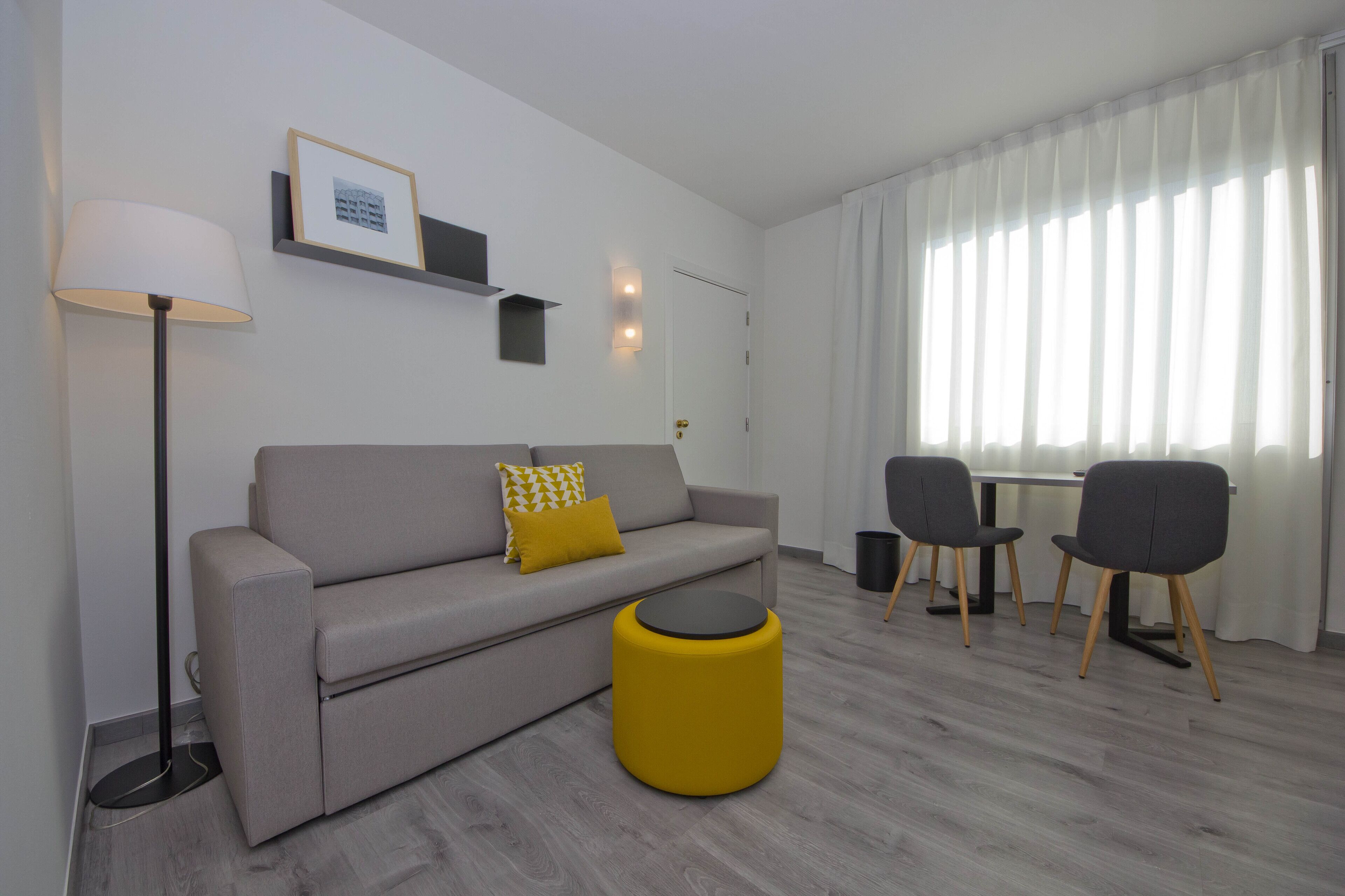Una sala de estar moderna con un sofá, un reposapiés amarillo, una lámpara de pie y un comedor con sillas.