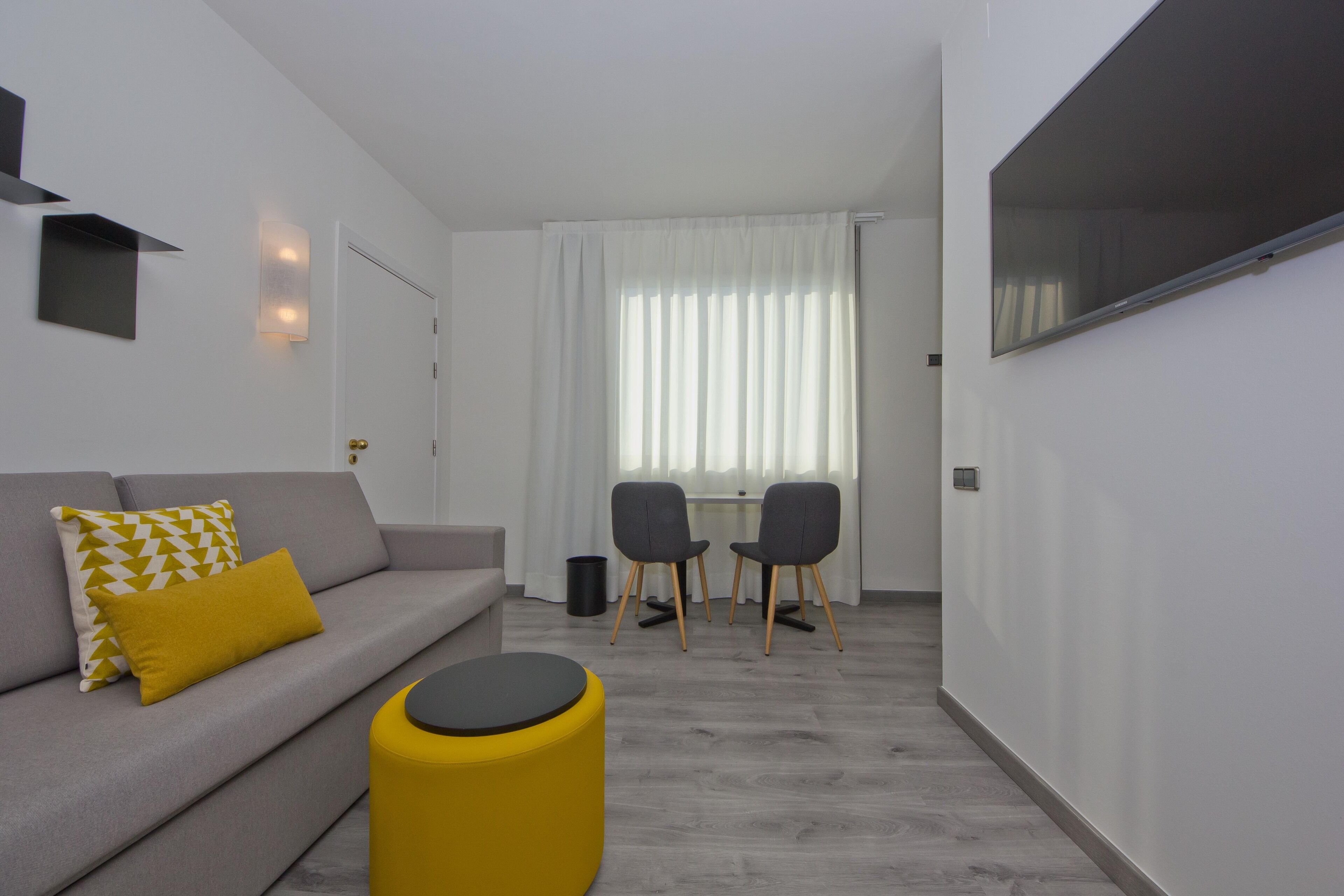 Una sala moderna con un sofá gris, almohadones blancos y amarillos estampados, un reposapiés amarillo y un comedor con dos sillas grises y una mesa.