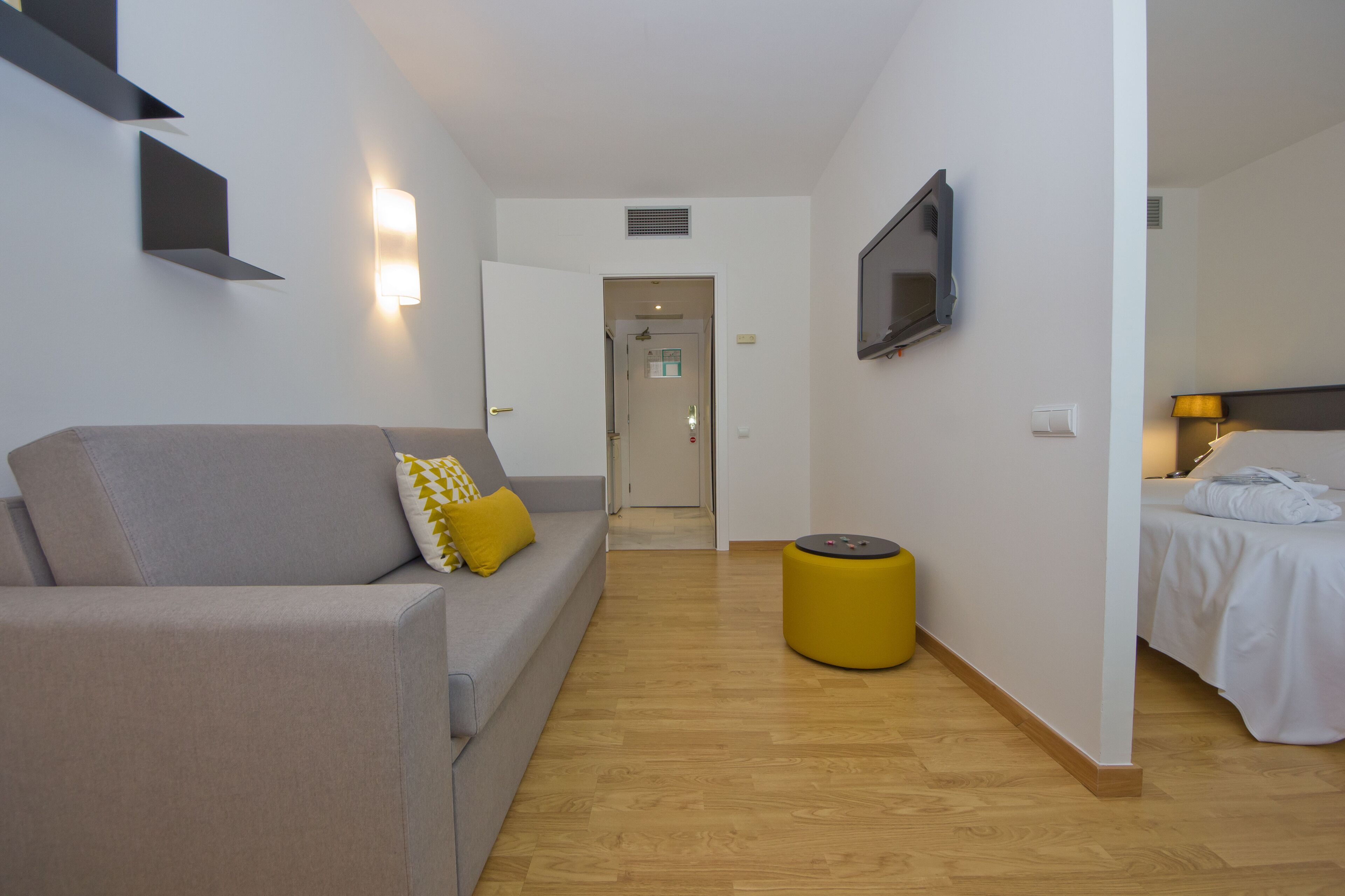 A modern hotel room with a grey sofa, a yellow ottoman, a bed with white linens, and a flat-screen TV mounted on the wall.