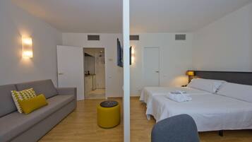 Una habitación de hotel moderna con un sofá gris, una cama con ropa de cama blanca, un pequeño otoman amarillo y suelo de madera.