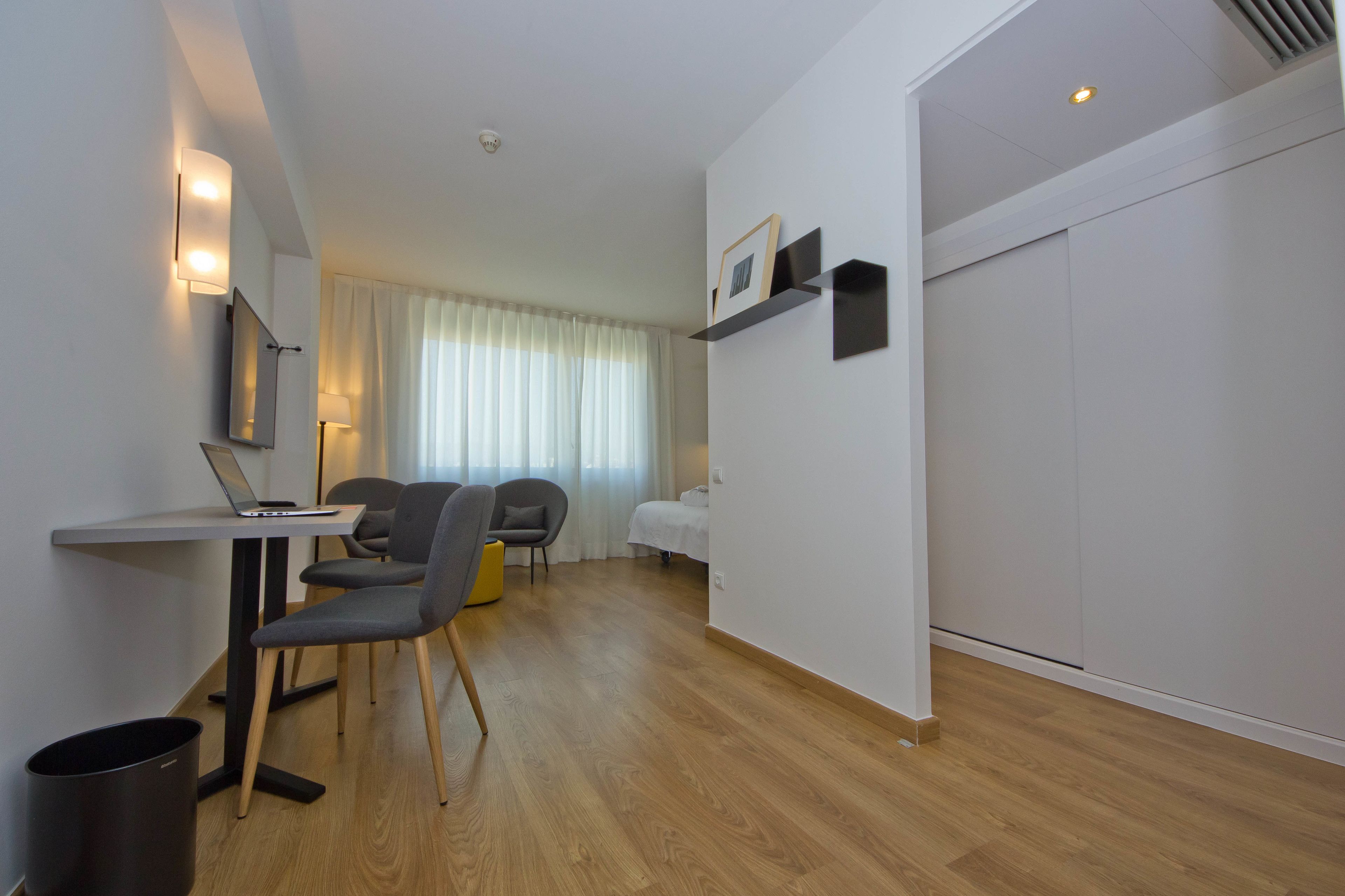 Una habitación de hotel moderna con piso de madera, un escritorio con una computadora portátil, dos sillas, una cama con cubrecama blanco, una mesita de noche y un estante de pared con un marco de fotos.