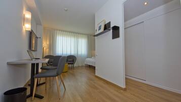 Una habitación de hotel moderna con piso de madera, un escritorio con una computadora portátil, dos sillas, una cama con cubrecama blanco, una mesita de noche y un estante de pared con un marco de fotos.