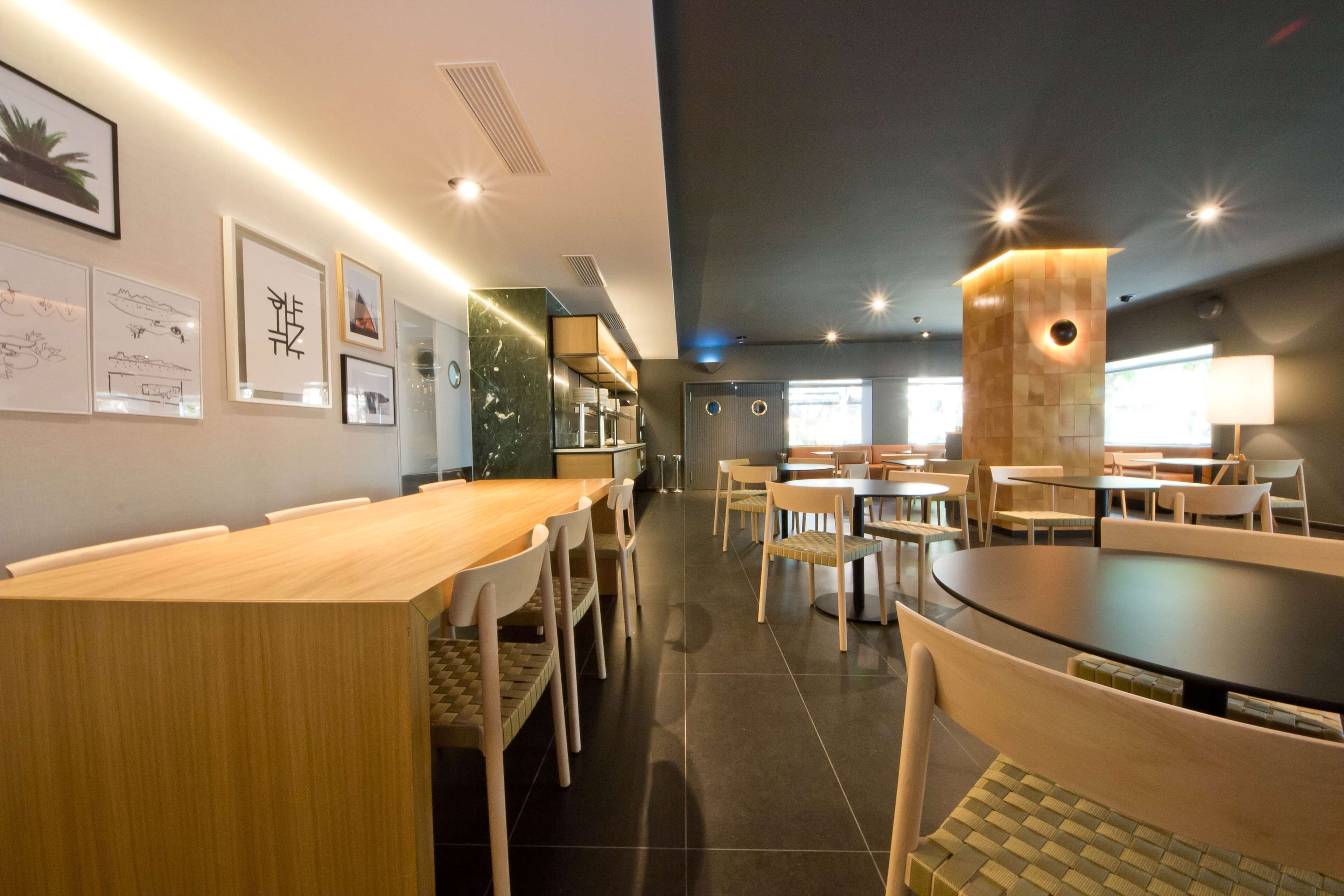 Un comedor moderno con mesas y sillas de madera, obras de arte en las paredes y una combinación de iluminación natural y artificial.