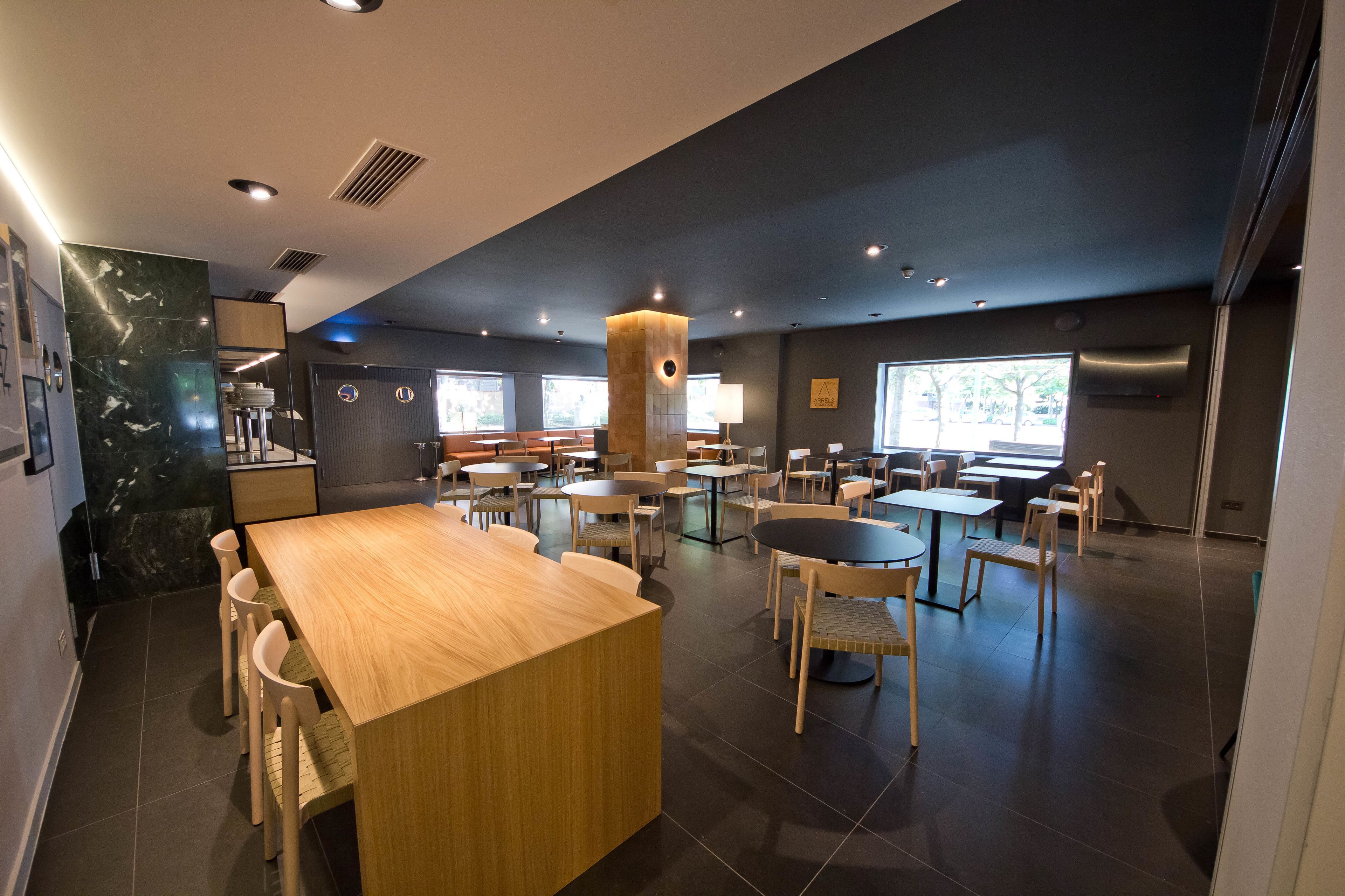Een modern restaurant met een centrale houten tafel, verschillende zithoeken en een groot raam dat voor natuurlijk licht zorgt.