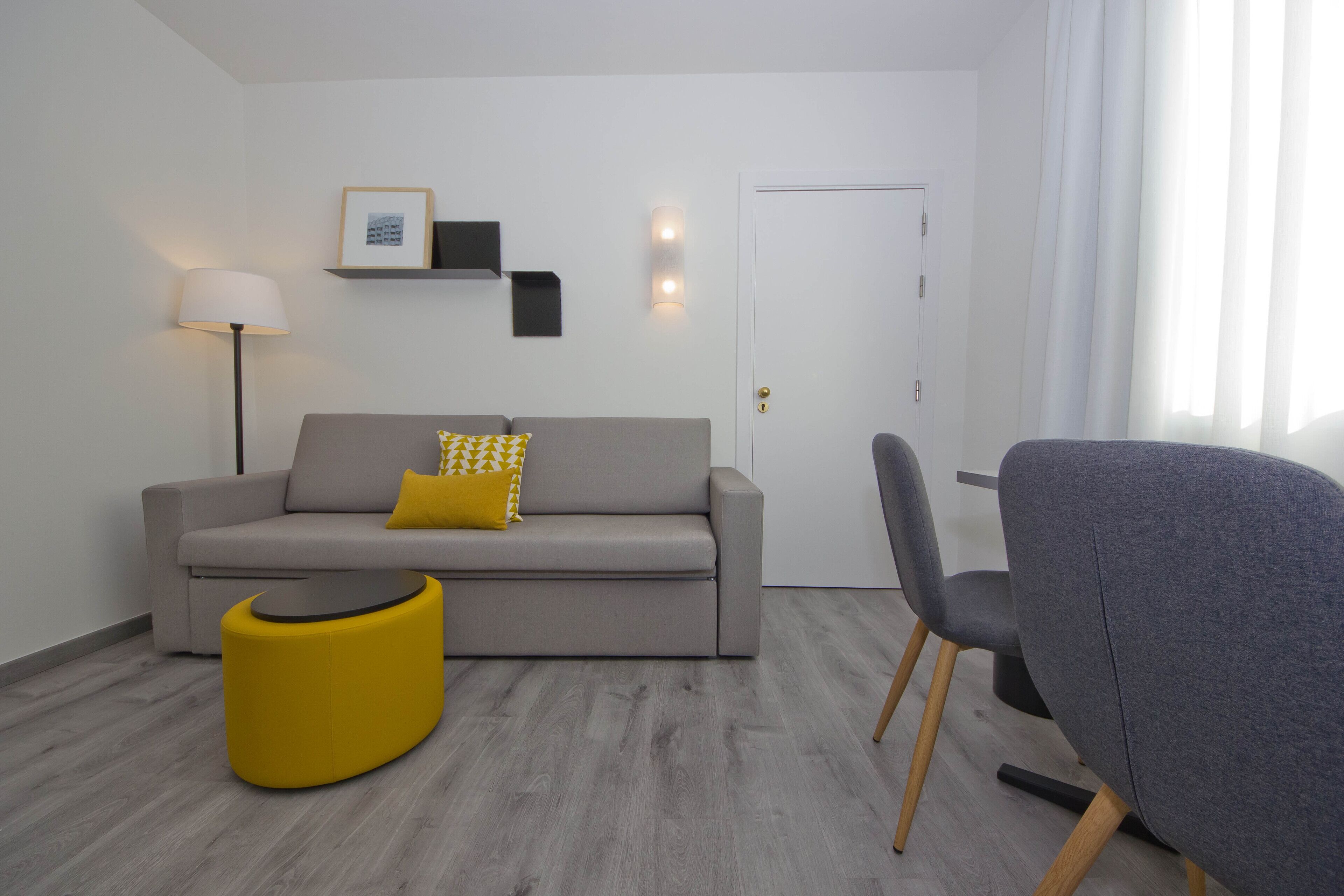 Una sala de estar moderna con un sofá gris, un reposapiés amarillo y una butaca gris. Hay una lámpara, un cuadro enmarcado y una lámpara de pared.
