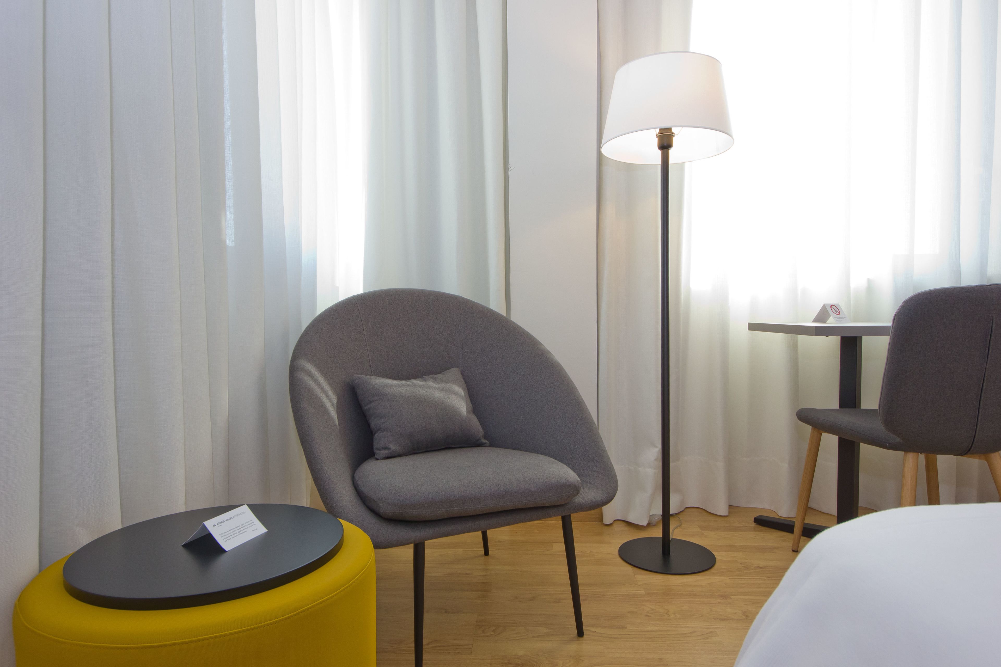 Una camera d'albergo con una poltrona grigia, un poggiapiedi giallo, una lampada da terra e un letto.