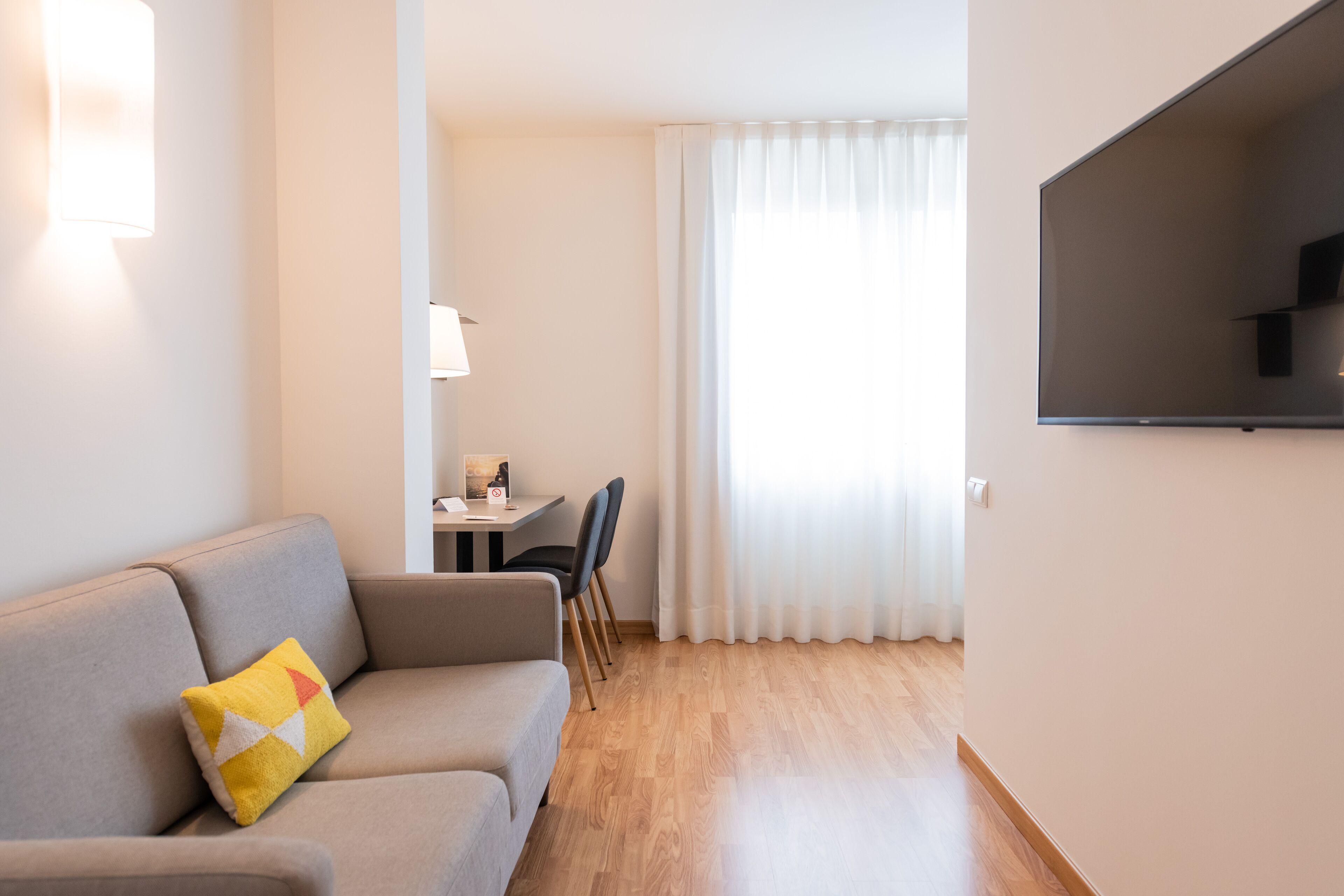 Una sala moderna con sofá, televisor de pantalla plana montado en la pared, zona de comedor con mesa y sillas, y un amplio ventanal con cortinas translúcidas.