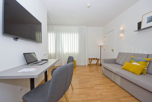 A modern living room with a grey sofa, a flat-screen TV mounted on the wall, a wooden floor, and a small table with a lamp. - Aparthotel Atenea Valles (Granollers)