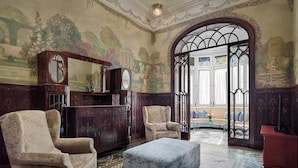 Una stanza con due poltrone, un poggiapiedi, un mobile in legno e un'ampia porta ad arco che dà su un balcone con vista.