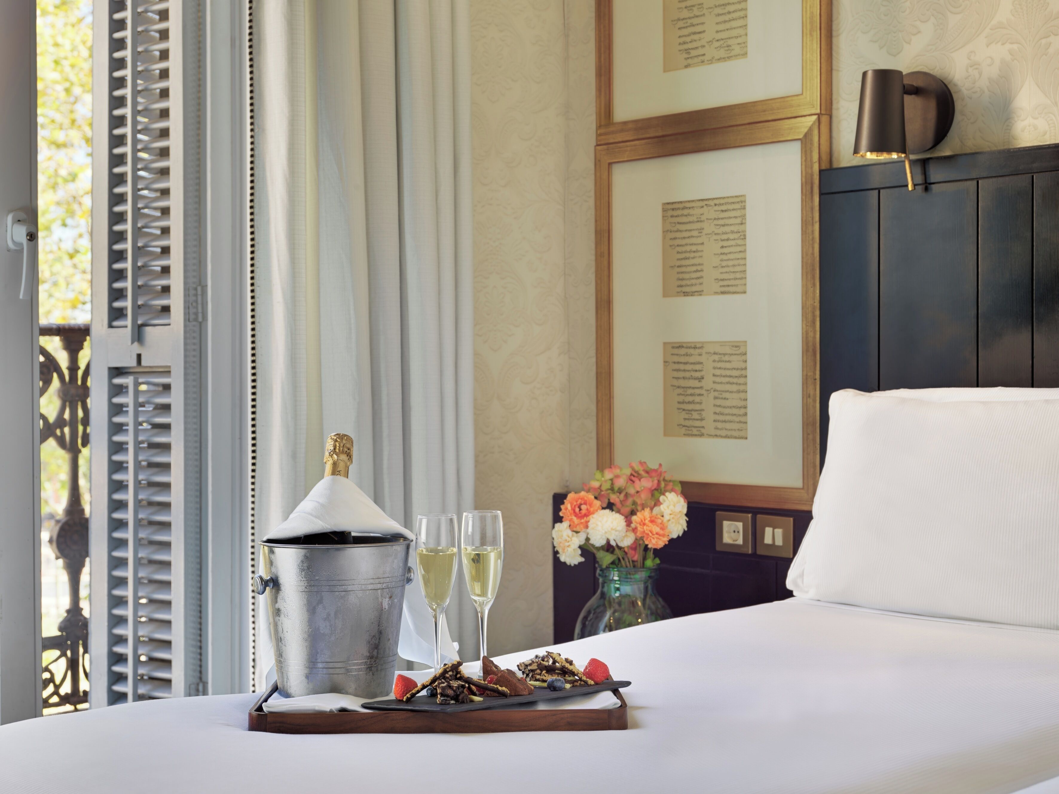 A hotel room with a bed, a tray with a champagne bucket, glasses, and a floral arrangement.