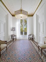 A room with a geometric patterned floor, two wooden chairs, a floor lamp, and a large window with a view of trees.