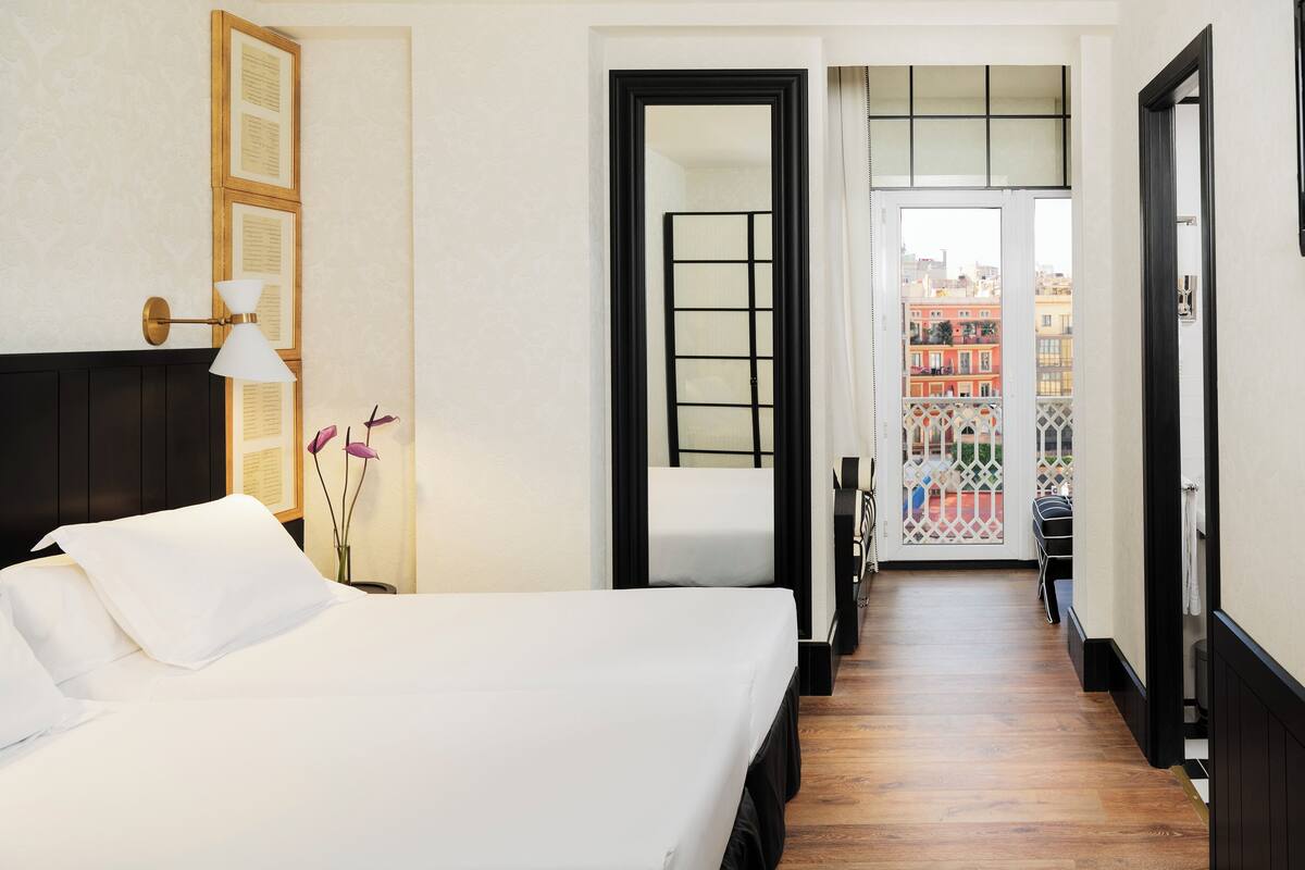 Une chambre d’hôtel avec un lit, un bureau et une vue sur la ville, accessible par une porte-fenêtre donnant sur un balcon.