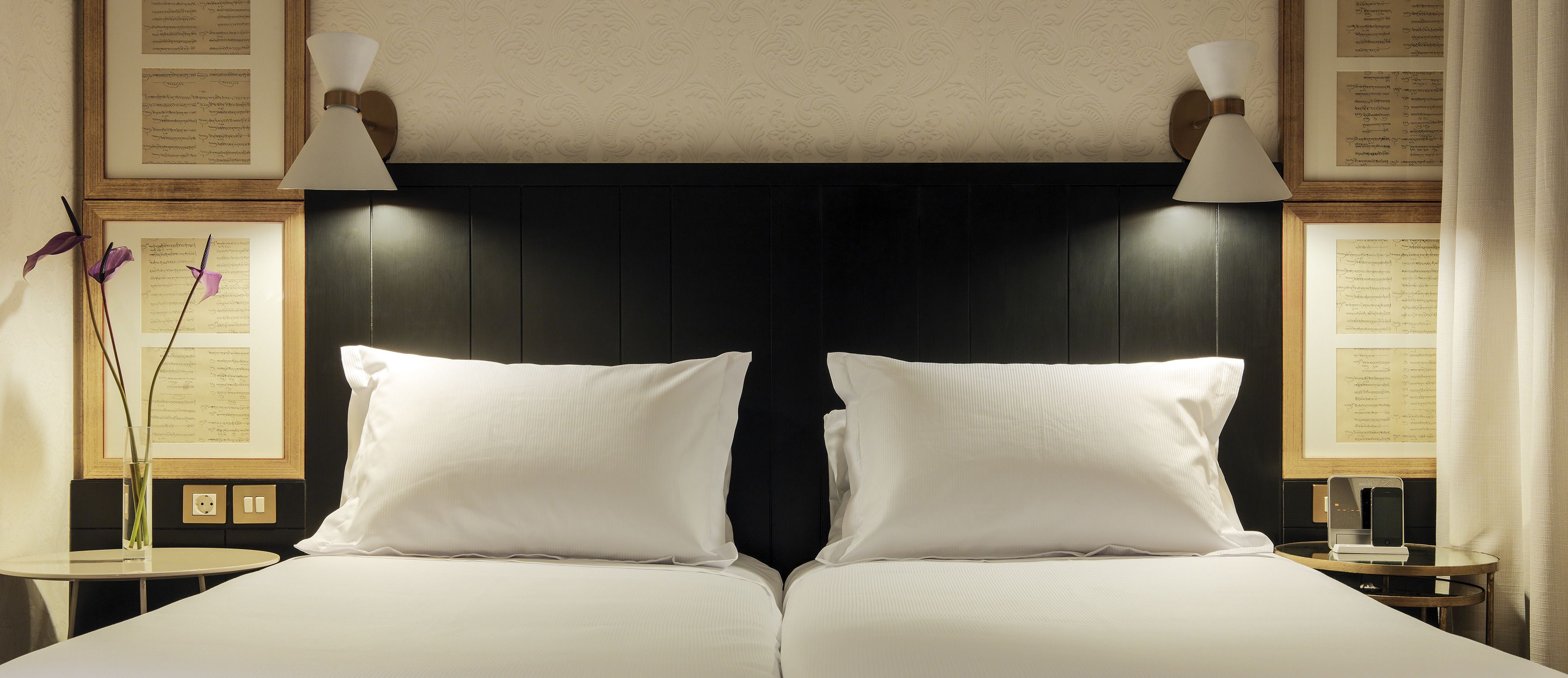 A double bed with white linens, two bedside tables with lamps, and framed documents on the walls.