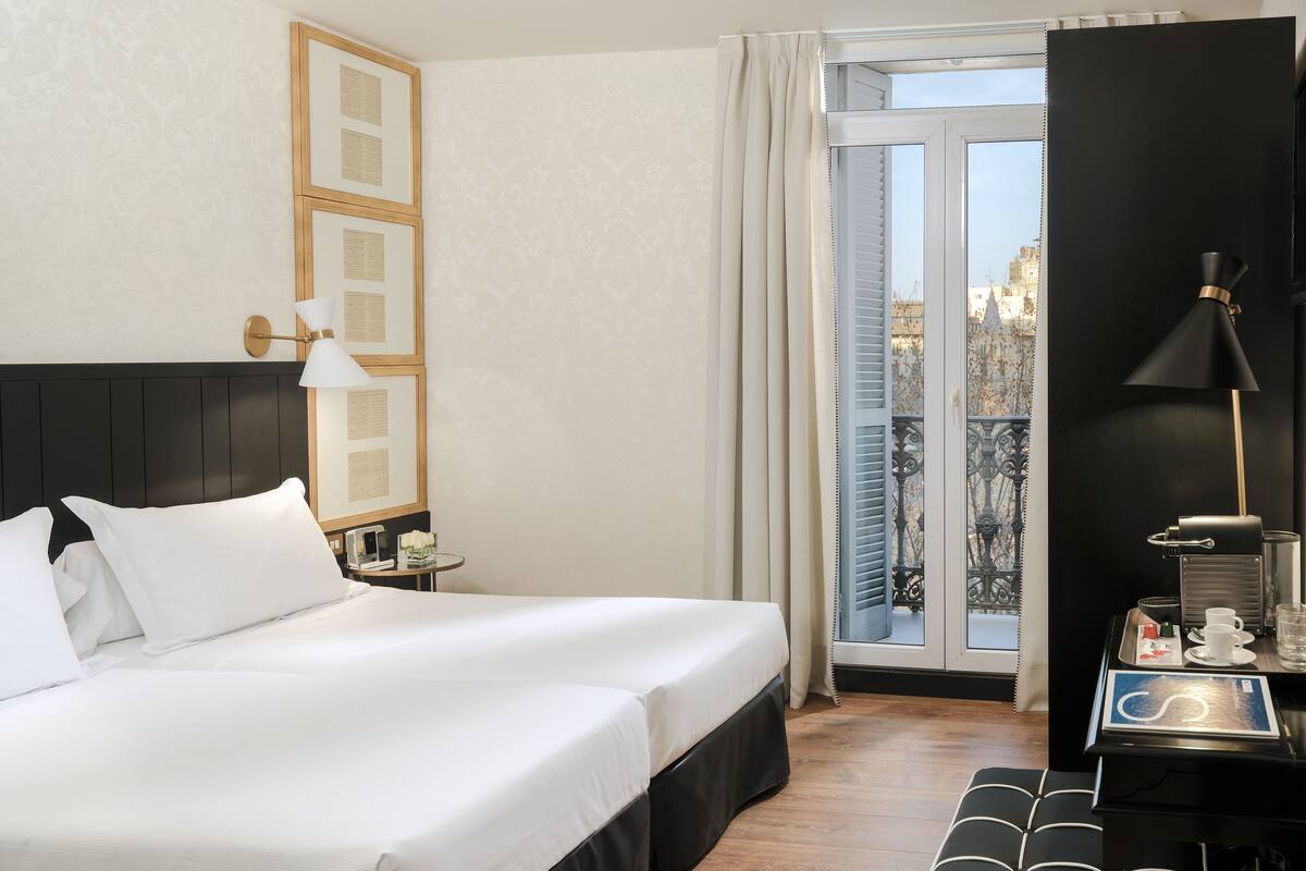 Une chambre d’hôtel avec un grand lit, un bureau avec une lampe, une vue sur la ville par la fenêtre et un balcon avec une balustrade.