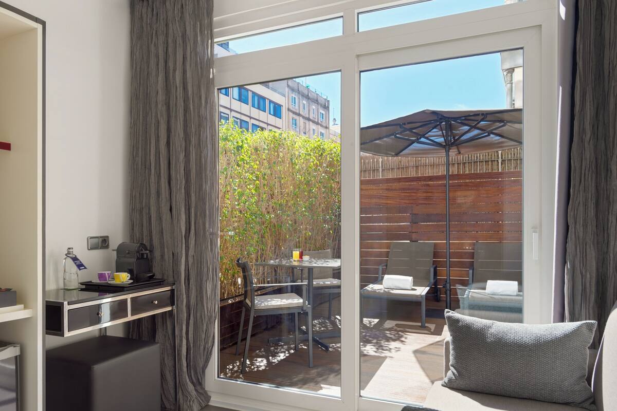 Une chambre moderne dotée d’une porte-fenêtre coulissante donnant sur un patio extérieur. À l’intérieur, on trouve un bureau avec une machine à café, une bouteille et une tasse. La chambre est également équipée d’un canapé et d’un fauteuil avec des coussins.