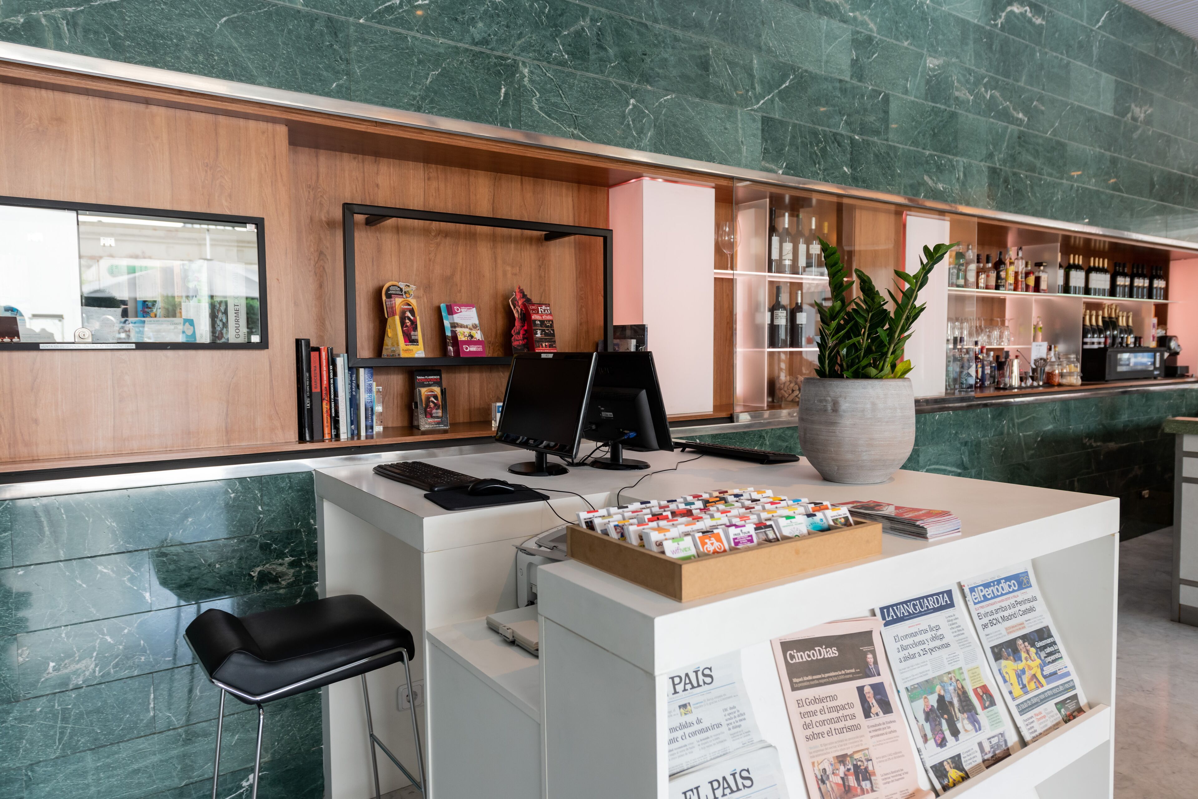 Un área de recepción con mostrador de mármol, una computadora, una planta y un mueble con revistas.
