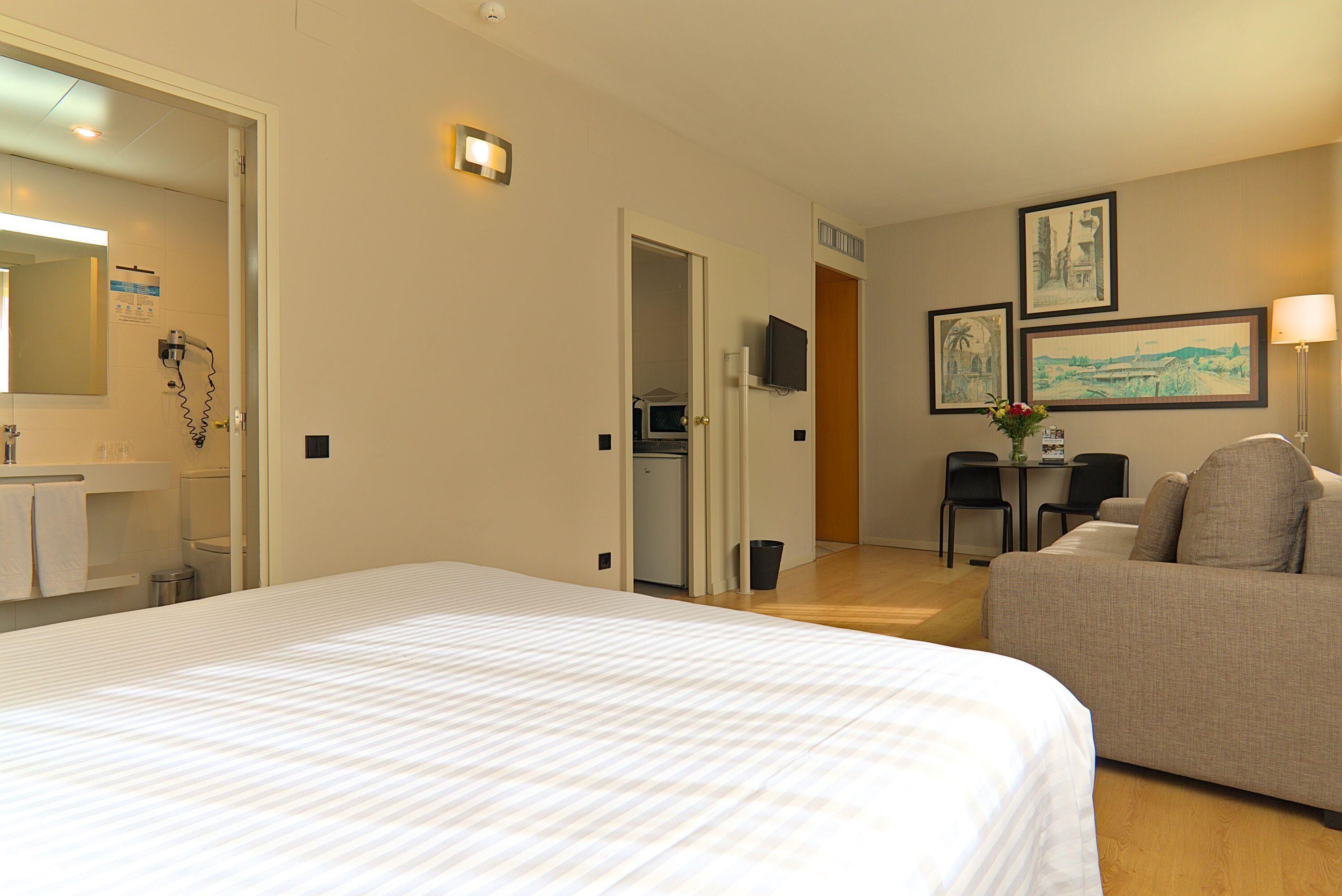 A hotel room with a large bed, a sofa, a desk with a chair, a TV, and artwork on the wall.