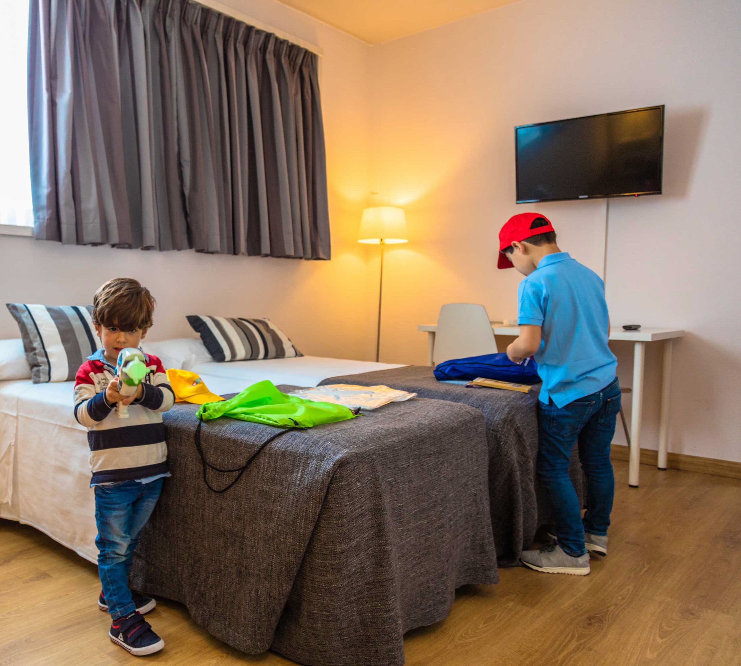 A hotel room with a bed, a desk, a television, and a child playing with toys.