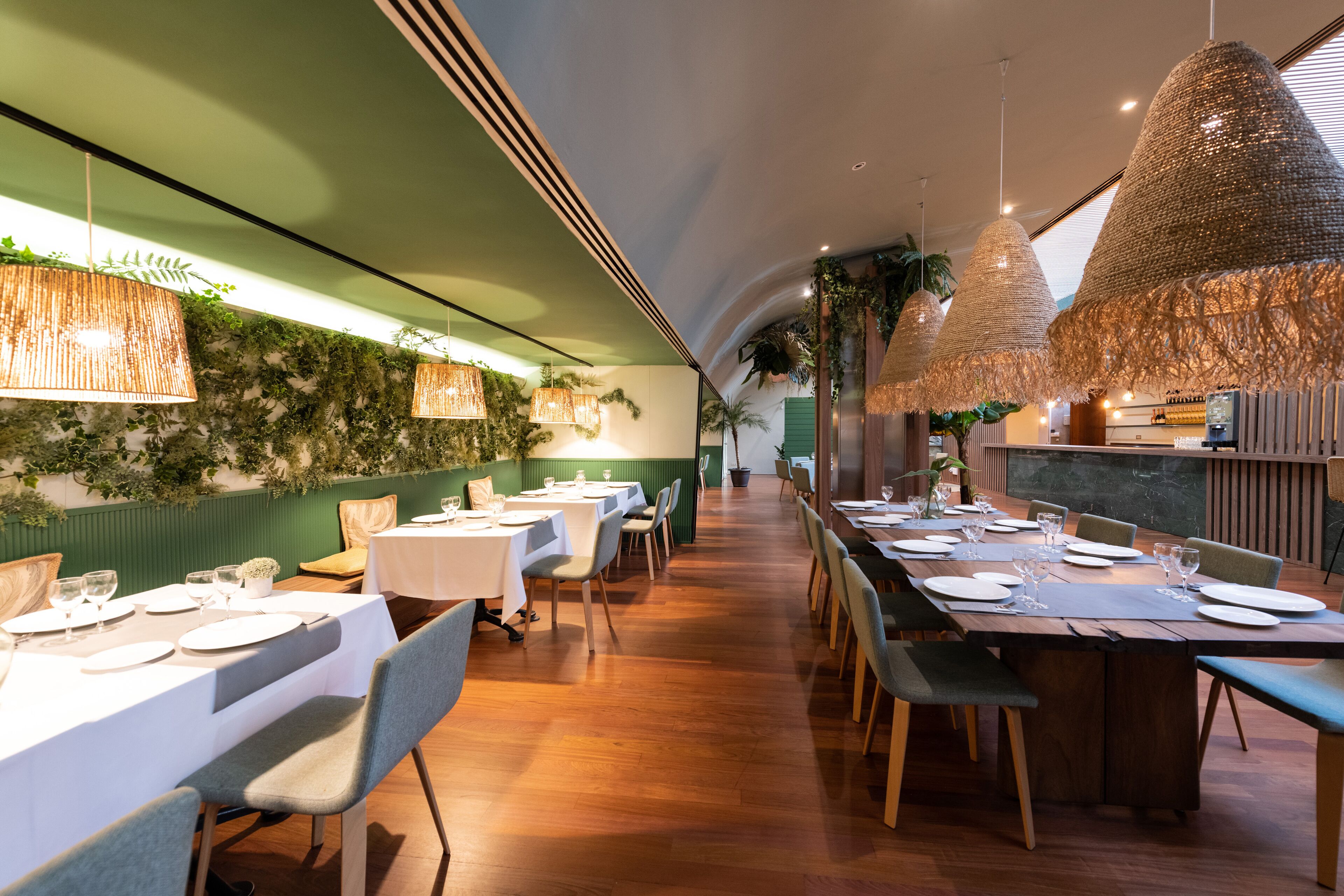 Een restaurant met lange houten tafels, witte tafellinnen en hangende geweven lampen.