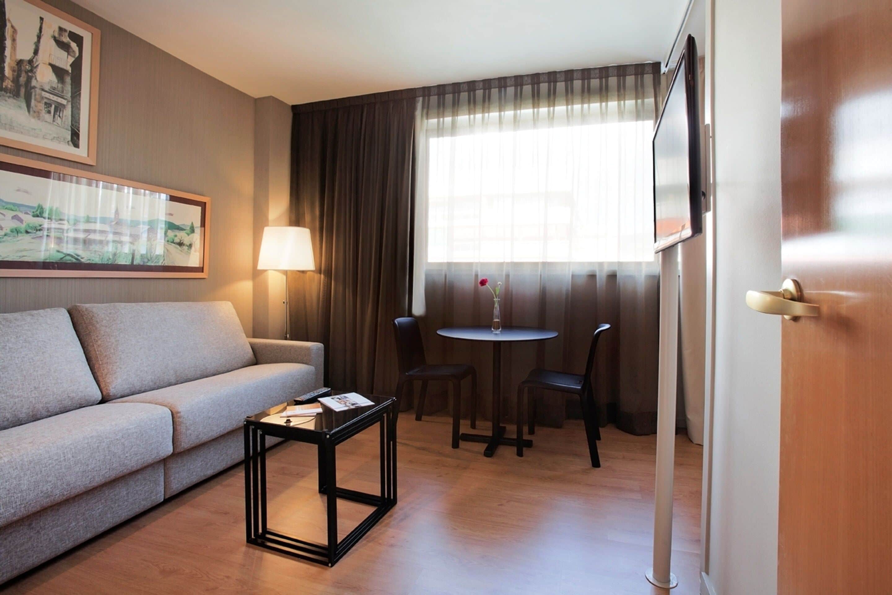 Une chambre d’hôtel avec un canapé, une petite table, une lampe, une télévision et une fenêtre avec des rideaux.