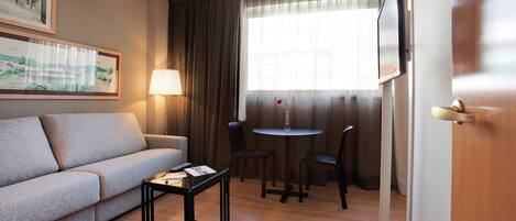 Une chambre d’hôtel avec un canapé, une petite table, une lampe, une télévision et une fenêtre avec des rideaux.