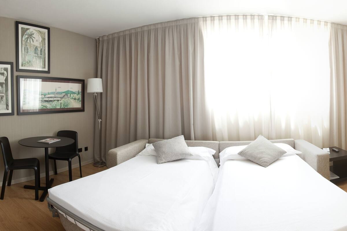 A hotel room with two beds, a desk, chairs, and a large window with curtains.
