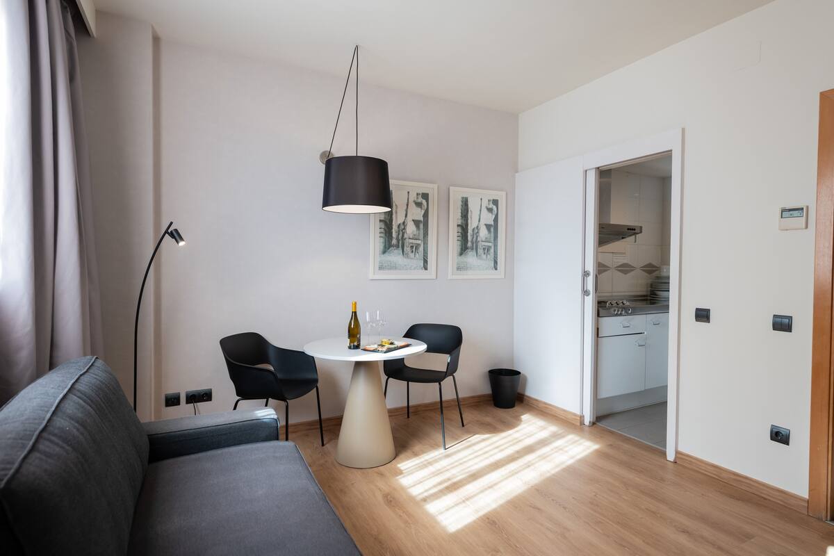 A modern living room with a sofa, a round table, and two chairs. There is a black pendant light, a floor lamp, and a door leading to a kitchen.