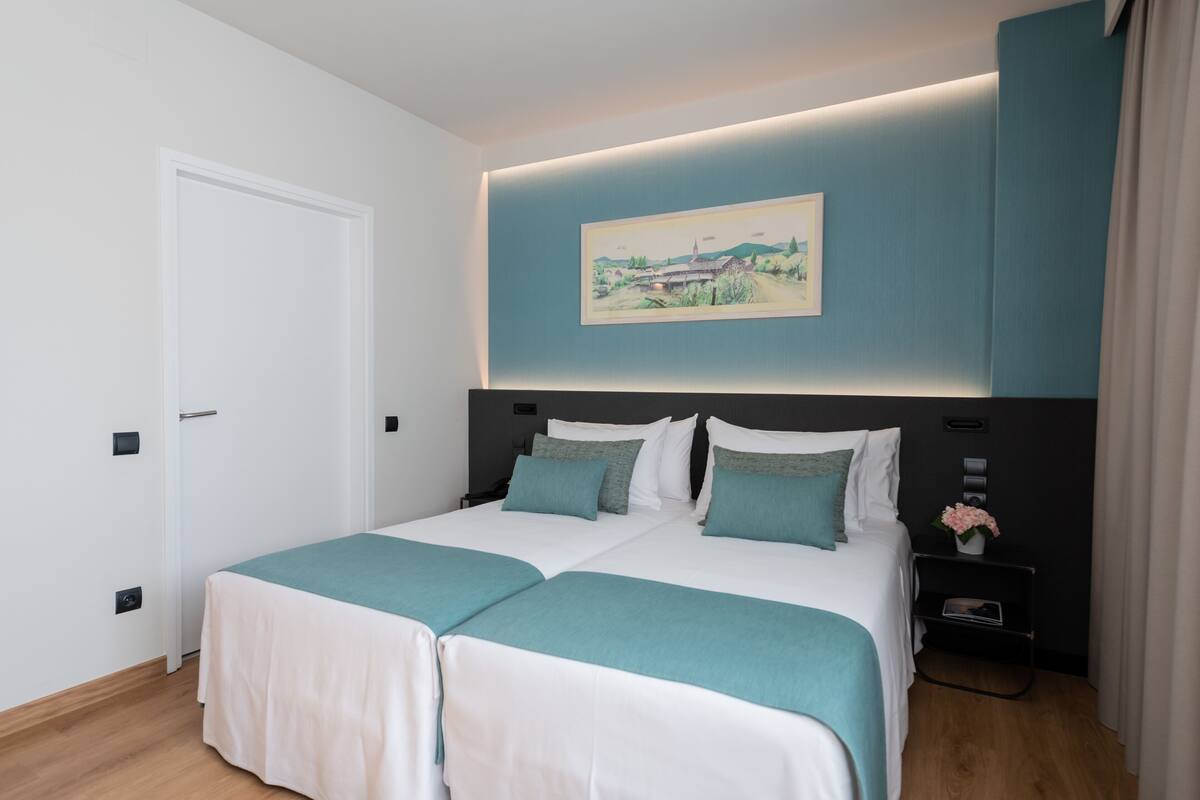 A double bed with a teal bedspread and white bedding, a nightstand with a vase of flowers, and a painting above the bed.