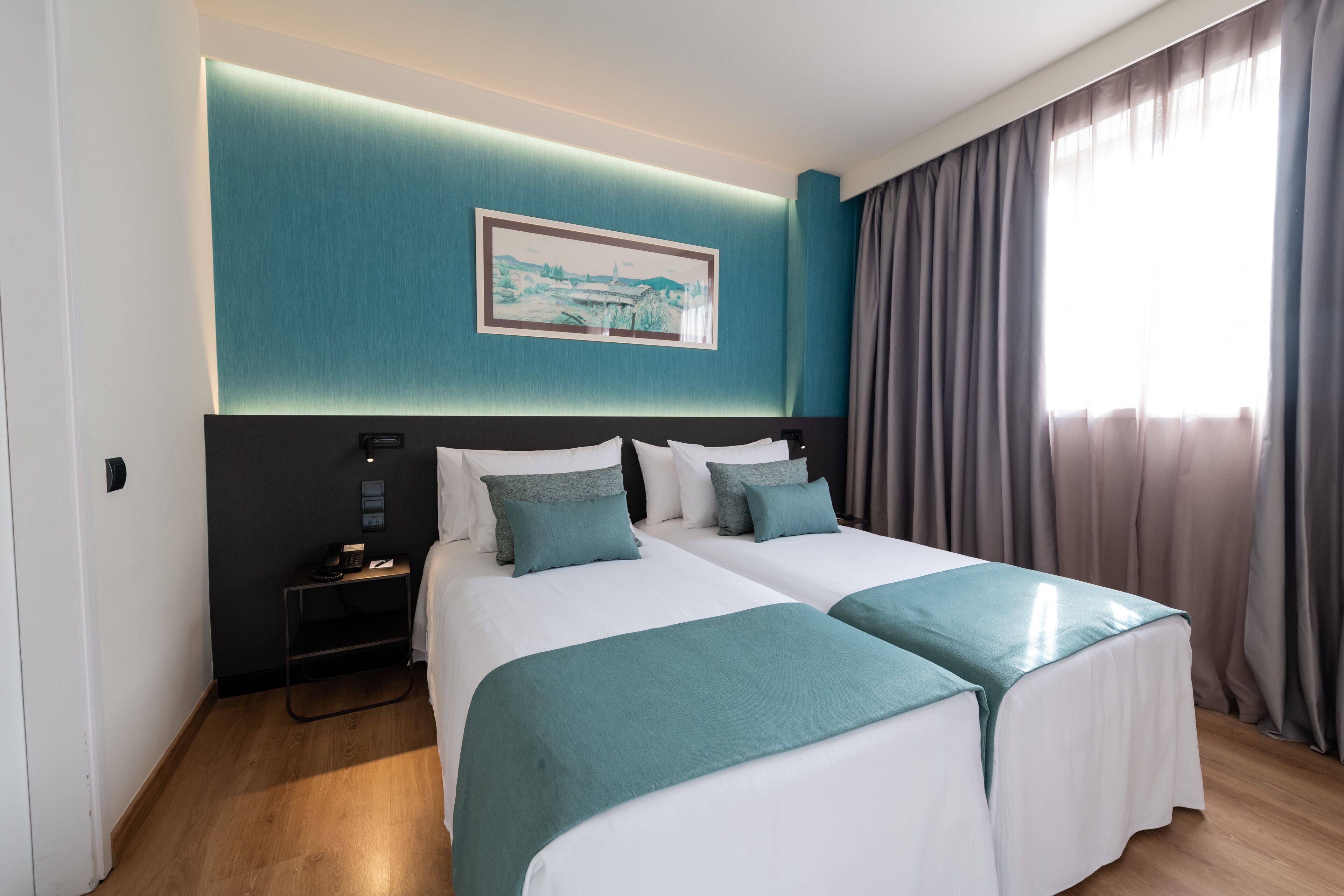 Habitación de hotel con dos camas, una mesita de noche, un cuadro en la pared y una ventana con cortinas.