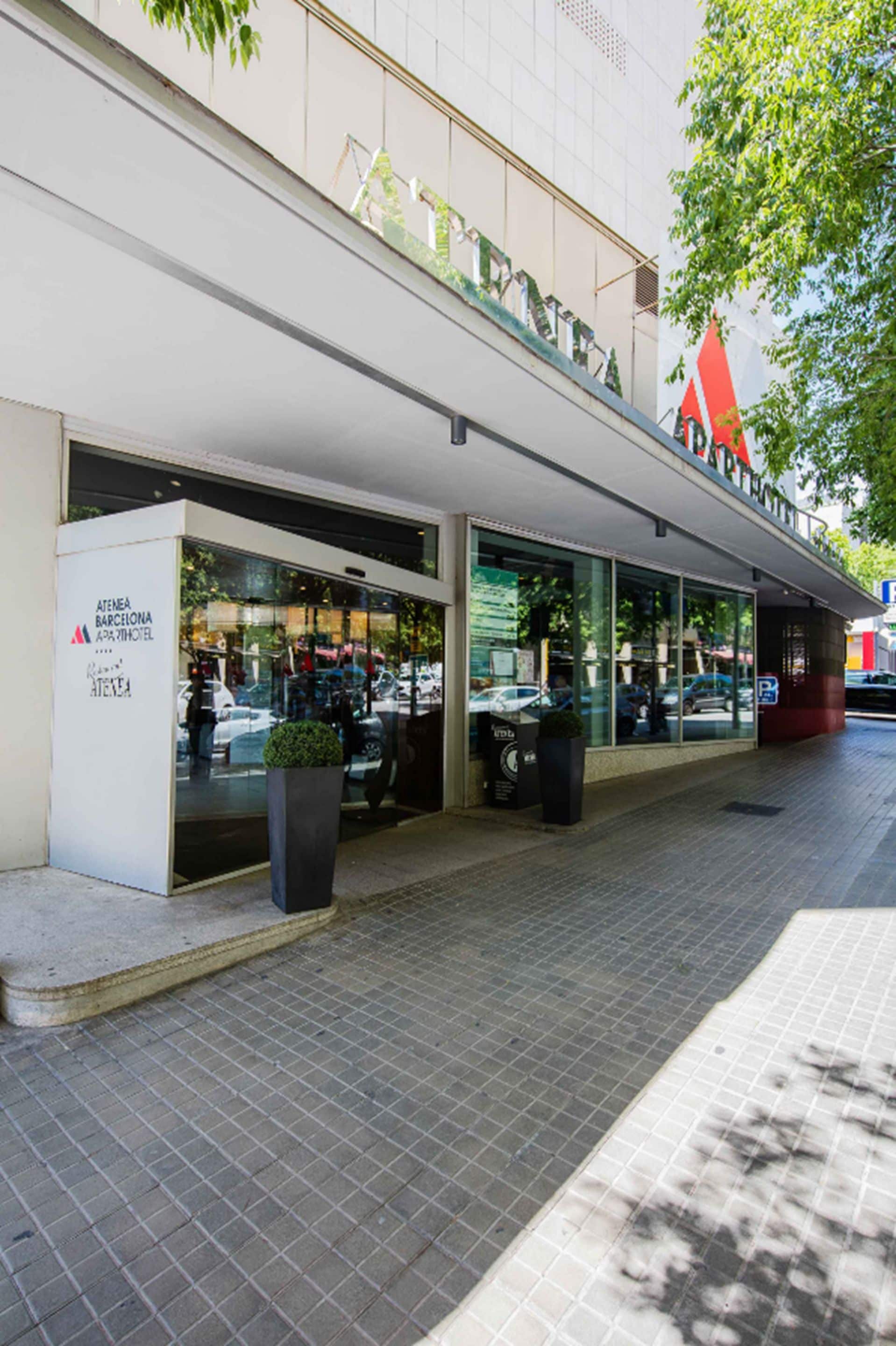 A modern building with a glass facade, a sign reading "Atenea Barcelona APOTHEC", and a parking area with a few cars.