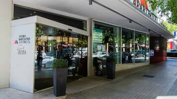 A modern building with a glass facade, a sign reading "Atenea Barcelona APOTHEC", and a parking area with a few cars.