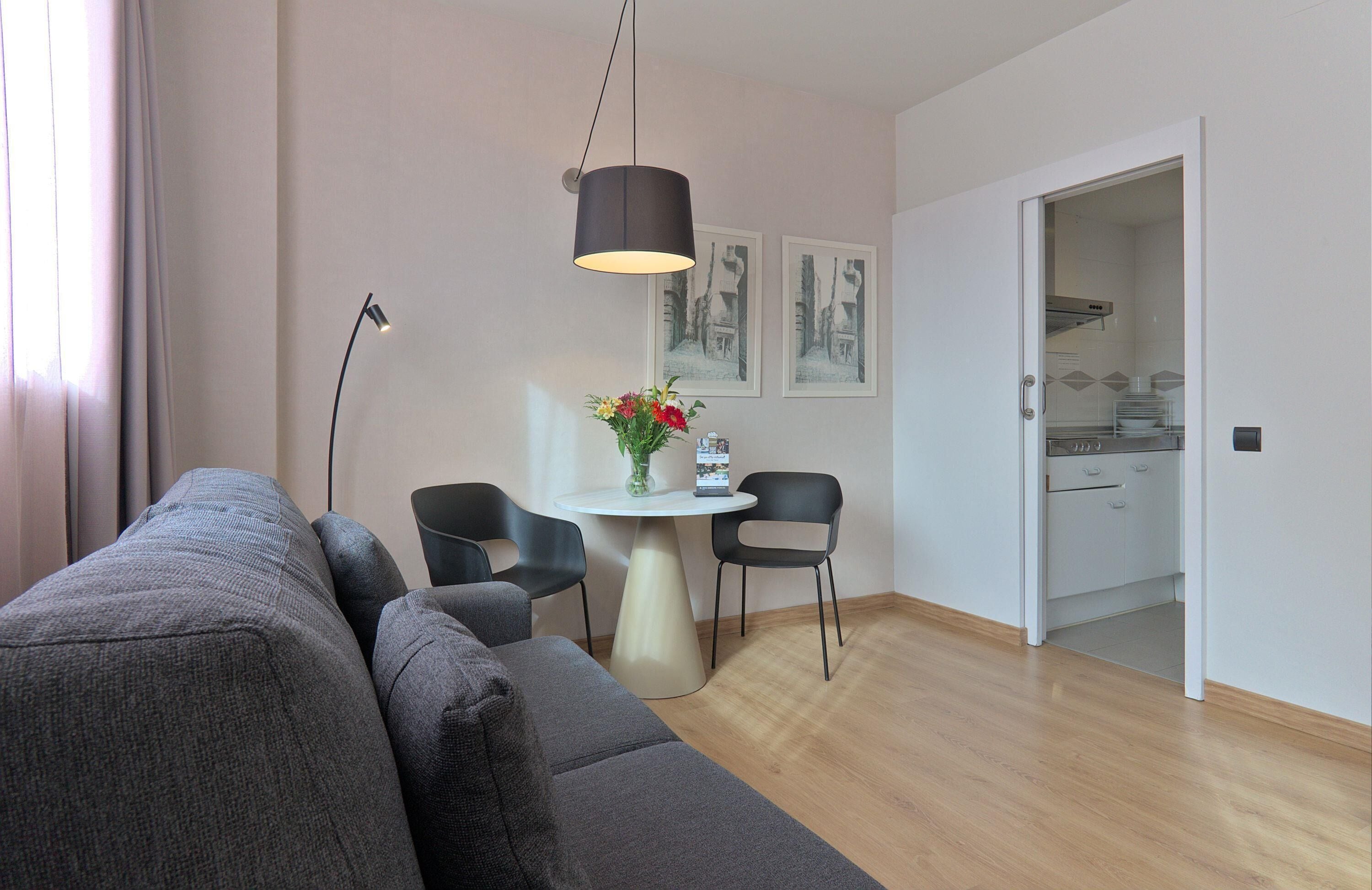 Una sala moderna con un sofá gris, una mesa redonda con sillas, un jarrón con flores y una cocina visible a través de una puerta abierta.