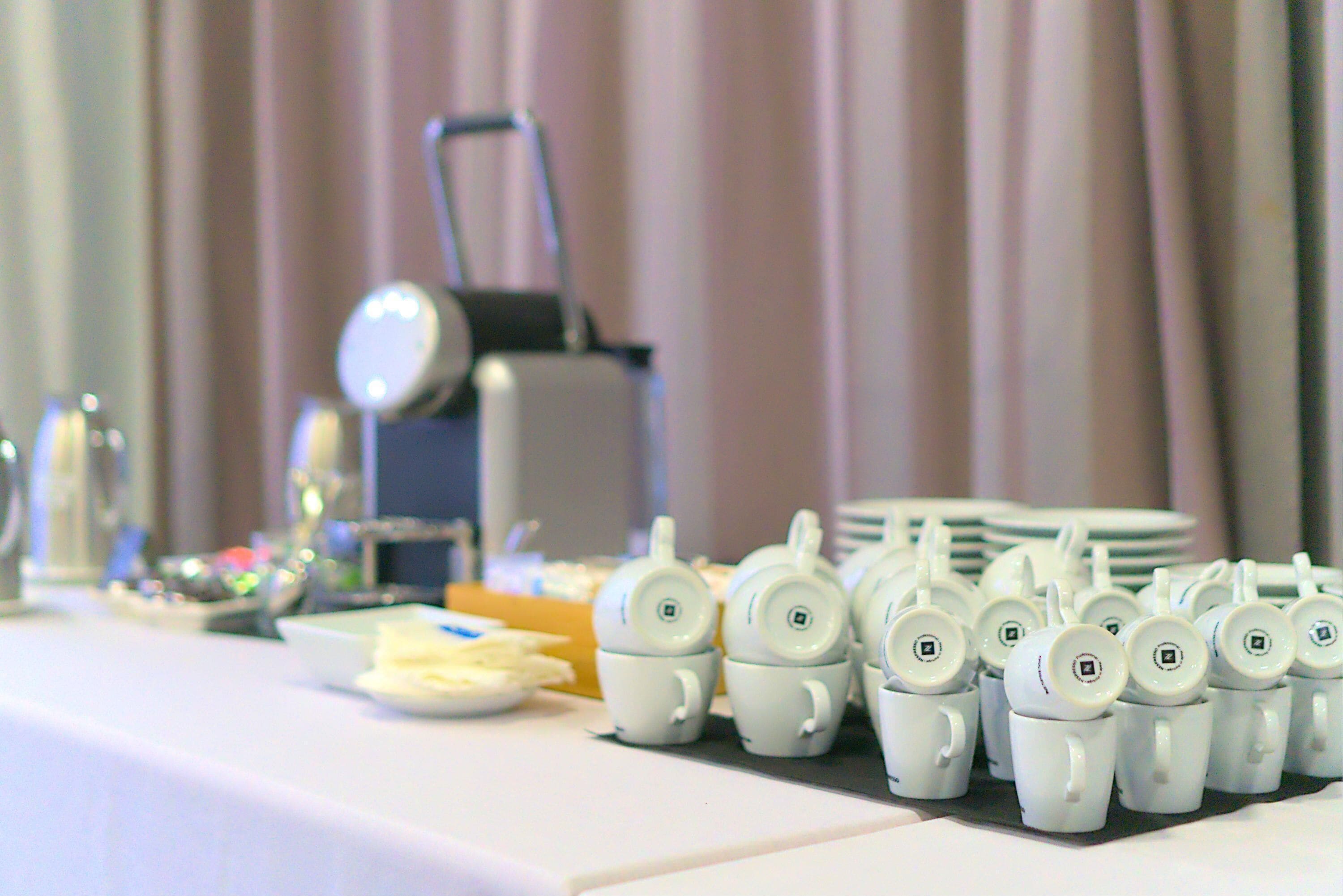 A table with multiple white teacups and plates, a small teapot, and a stack of plates in the background.