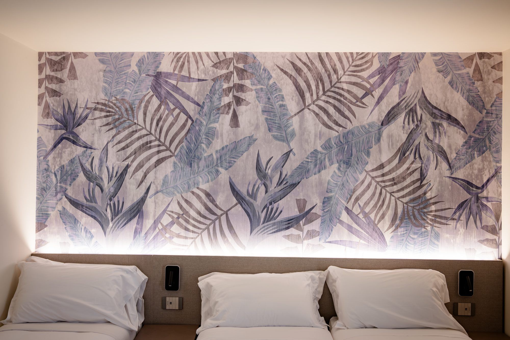 A hotel room with a large wall tapestry featuring tropical leaves, two beds with white linens, and a window with blinds.