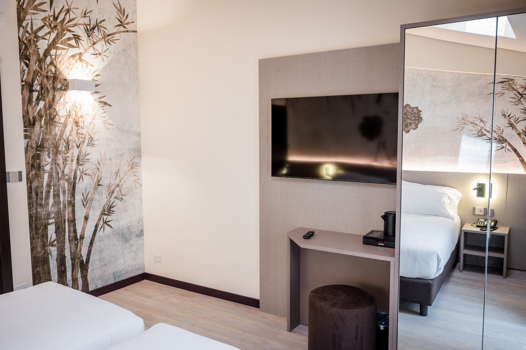 Quarto de hotel moderno com uma cama grande, uma mesa com computador, um espelho e TV na parede.