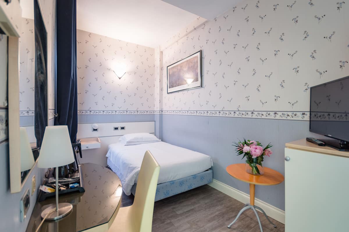 Una camera d'albergo con un letto, una scrivania con una lampada, una sedia, un piccolo tavolino con un vaso di fiori e una televisione.