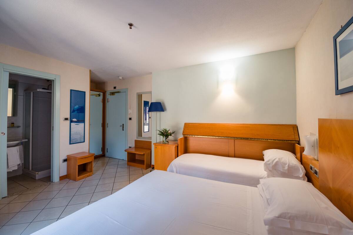 Una camera d'albergo con due letti, una doccia, una piccola scrivania e una lampada.