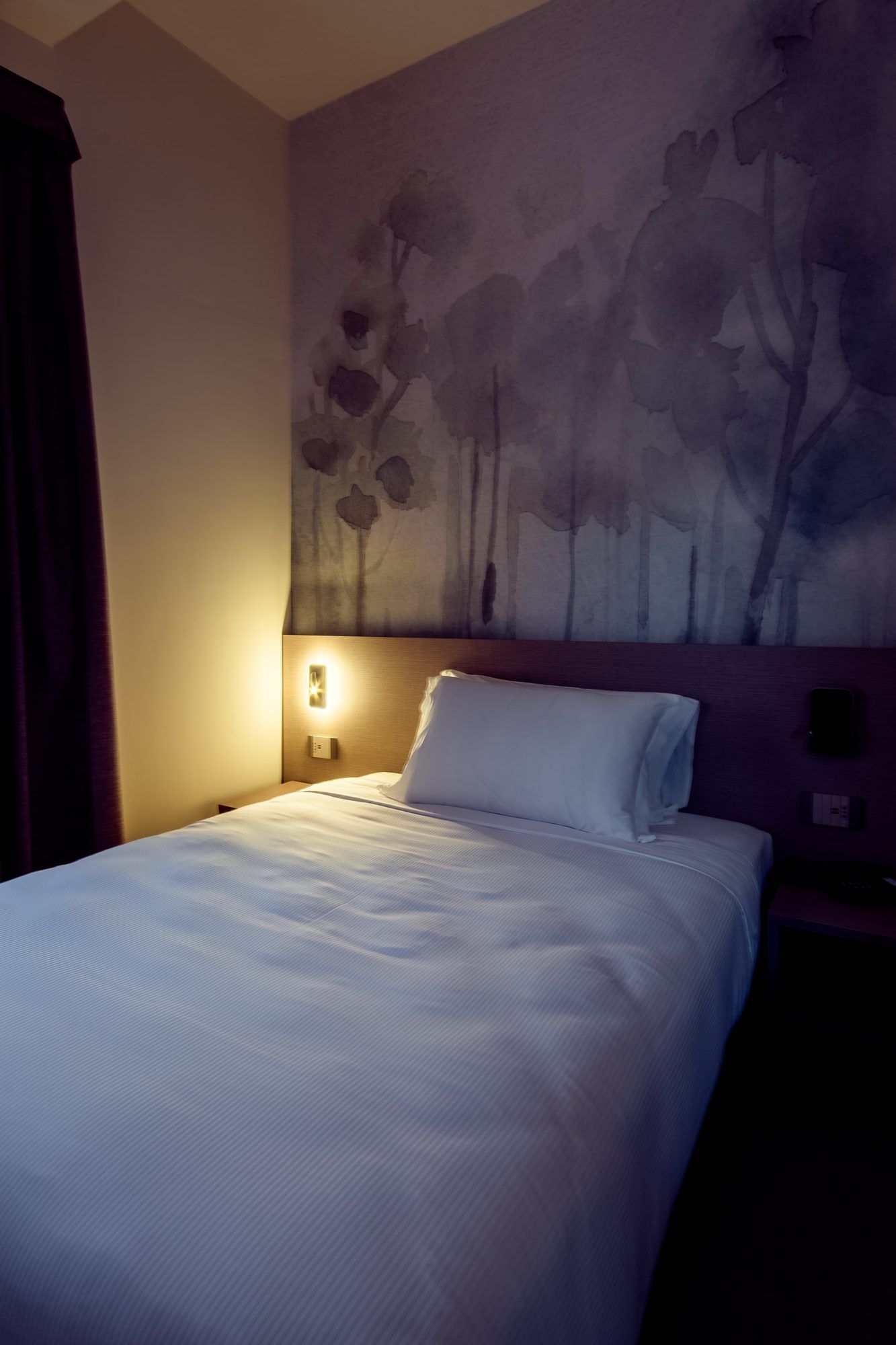 A hotel room with a bed, a wall-mounted light, and a painting.