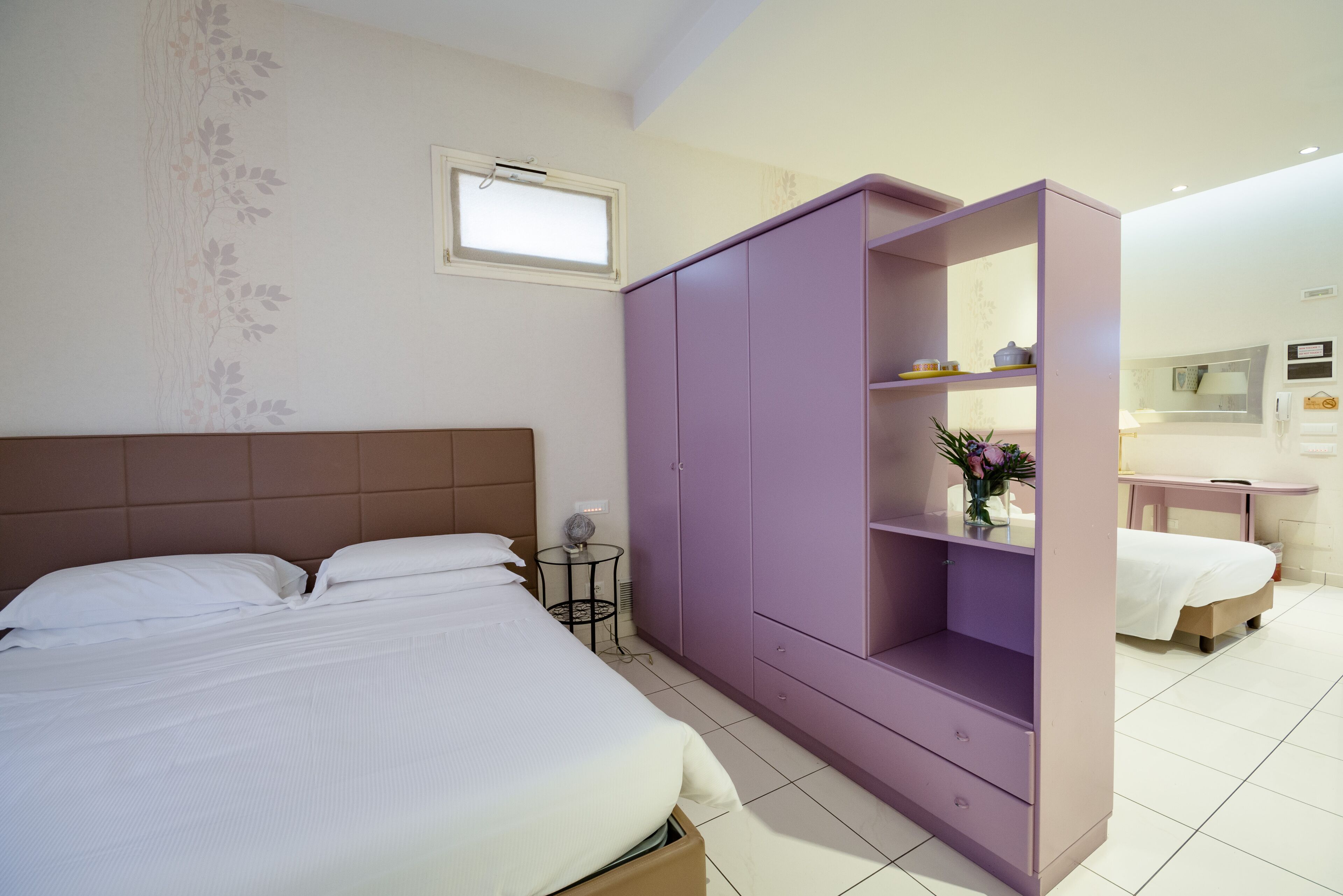 A bedroom with a bed, a purple wardrobe, a small table with a lamp, and a bathroom in the background.