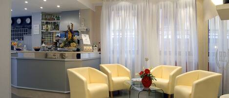 A modern hotel lobby with a reception desk, seating area with armchairs, a glass coffee table, and a large window with sheer curtains.