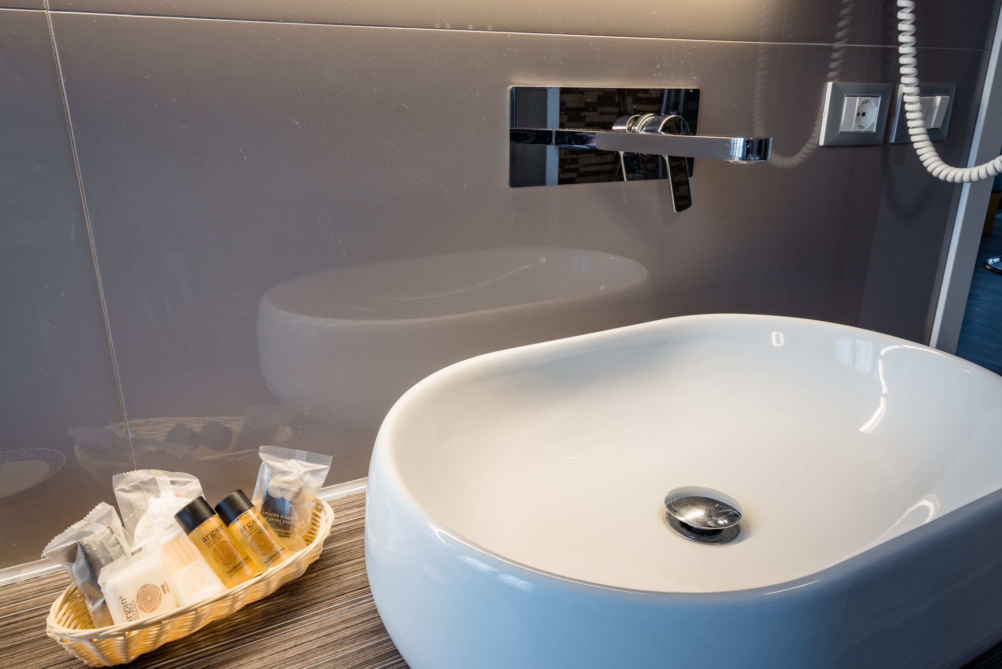 Un baño moderno con un lavamanos ovalado grande, un box de ducha de vidrio y una cesta de mimbre con artículos de aseo.